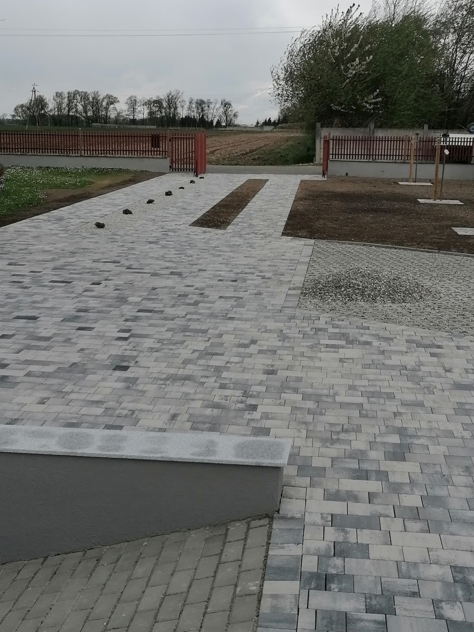 Ułożona kostka brukowa w odcieniach szarości tworząca ścieżkę i podjazd. W tle widoczne ogrodzenie i drzewa. Minimalistyczny styl, świeżo po realizacji.