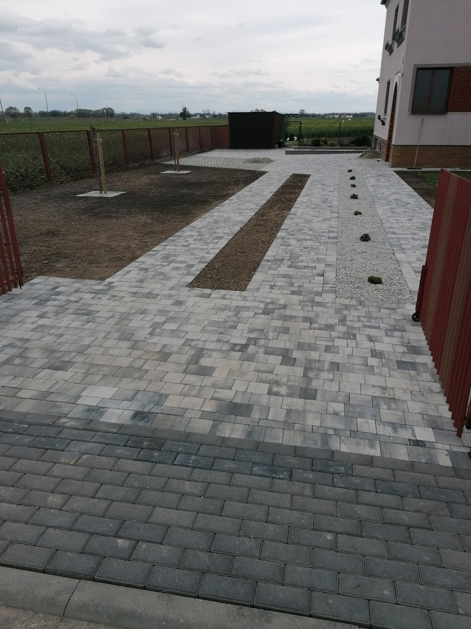Ułożona kostka brukowa w odcieniach szarości tworząca ścieżkę i podjazd do domu. Elementy dekoracyjne z kamienia i ziemi. Widok z góry na posesję.