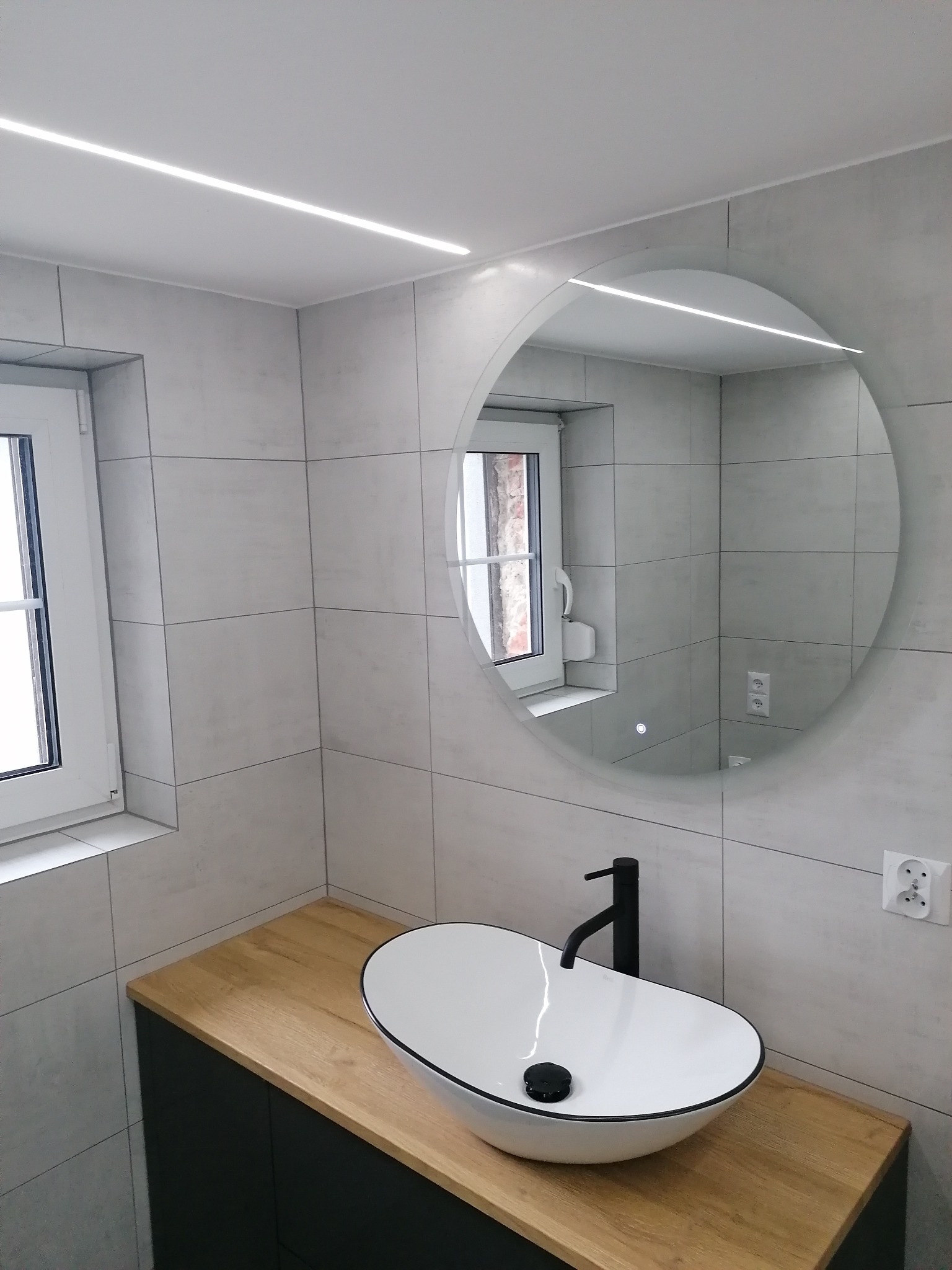Nowoczesna łazienka z owalną umywalką nablatową, drewnianym blatem i okrągłym lustrem z podświetleniem LED. Szare płytki na ścianach i sufitowe oświetlenie liniowe.