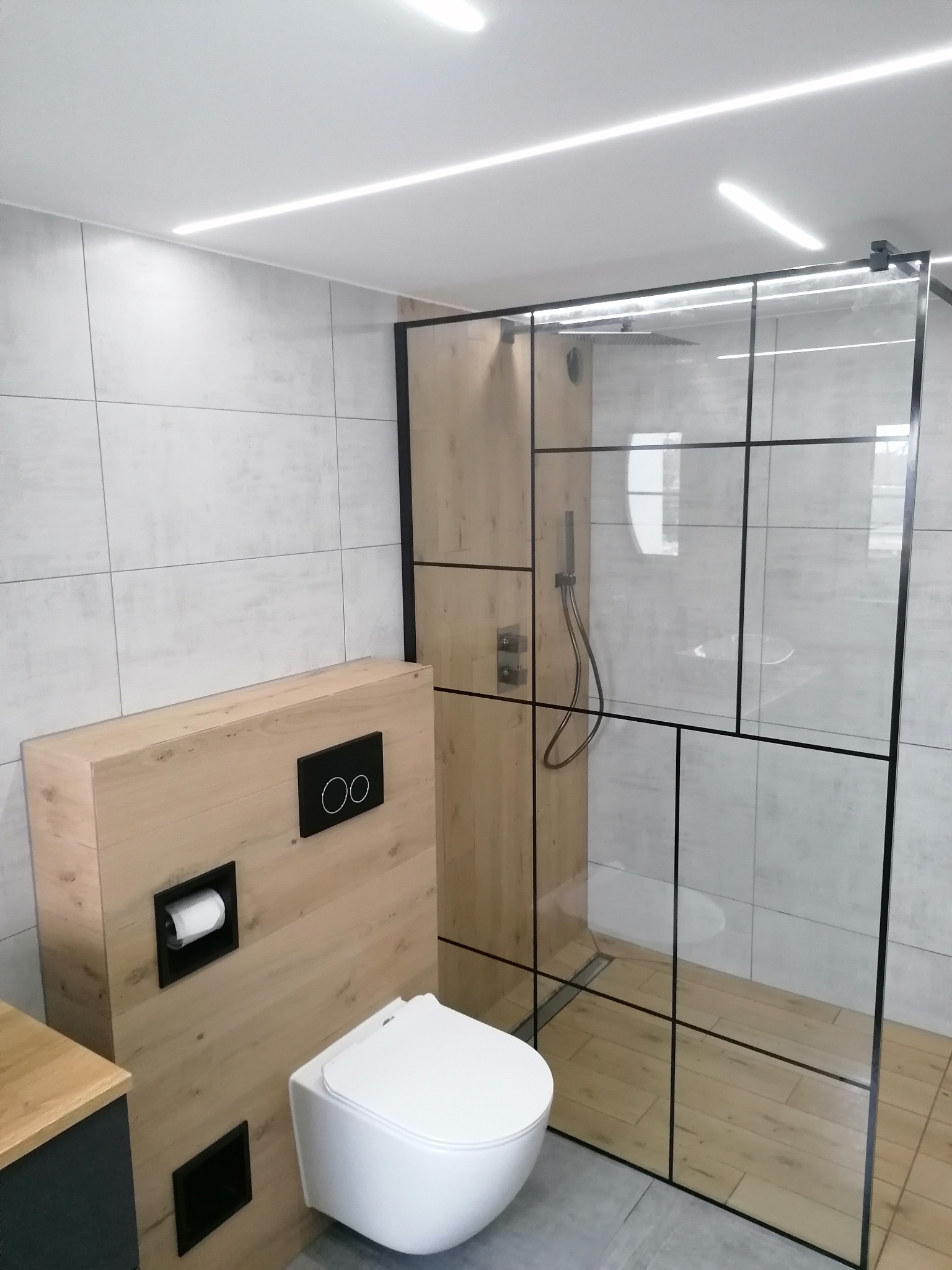 Nowoczesna łazienka z kabiną prysznicową typu walk-in, drewnopodobną zabudową WC i szarą płytką. Minimalistyczny design z czarnymi akcentami.
