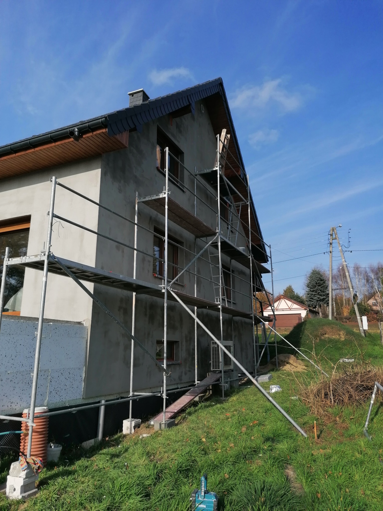 Dom w trakcie ocieplania elewacji, widoczne rusztowania, częściowo ocieplona ściana z szarym tynkiem, błękitne niebo w tle. Ujęcie z dołu, z prawej strony budynku.