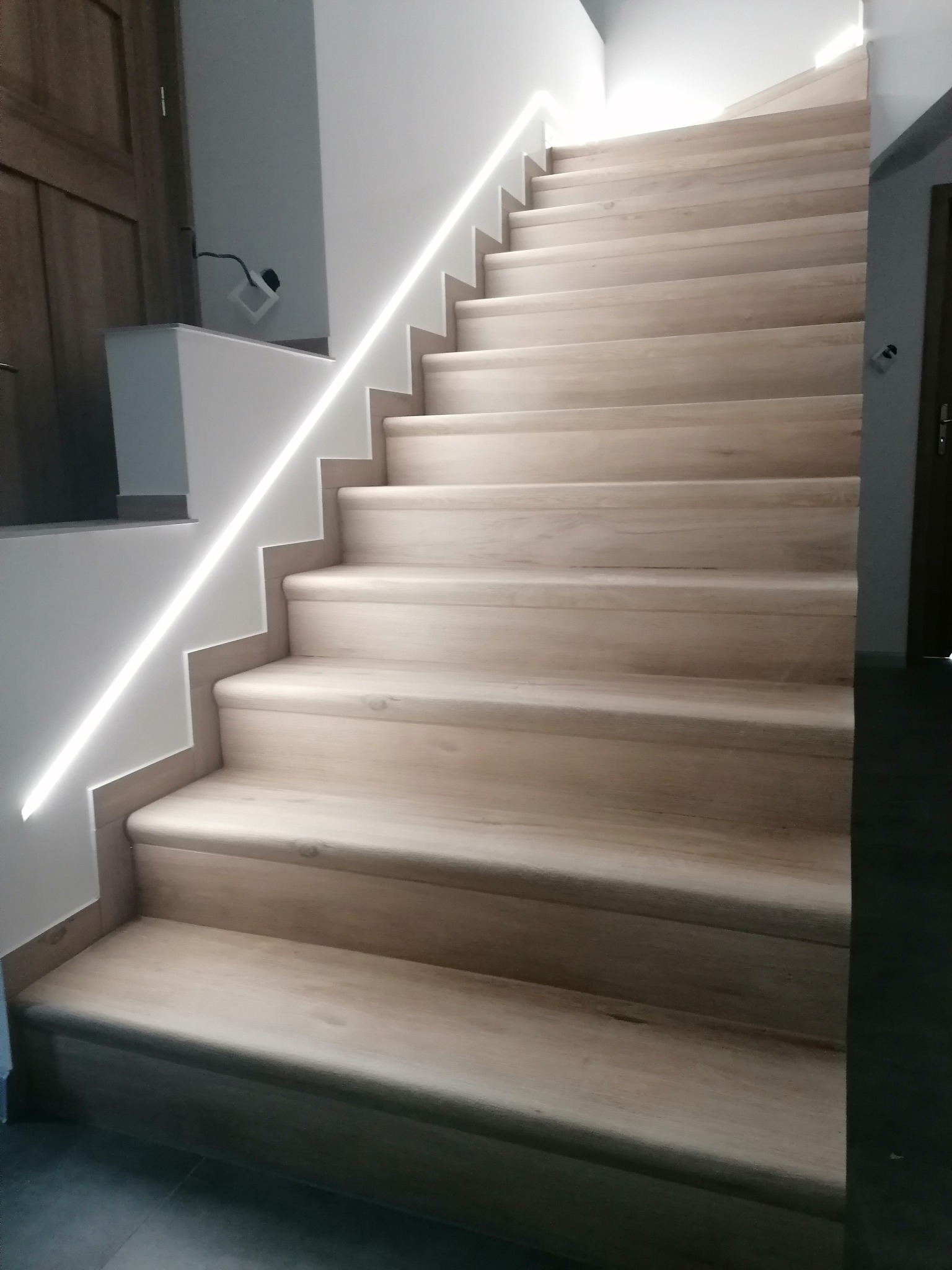 Jasne, drewniane schody z subtelnym oświetleniem LED wzdłuż ściany, tworzące nowoczesny i elegancki wygląd wnętrza. Widok z dołu, podkreślający detale wykończenia.