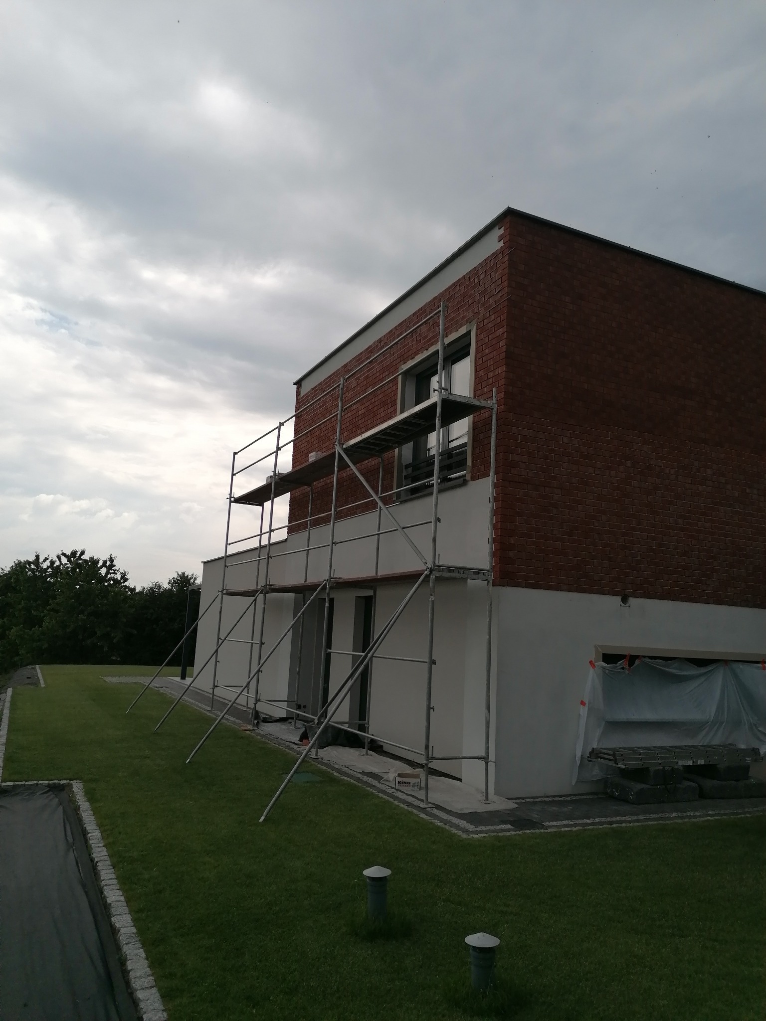 Dwukondygnacyjny dom z czerwoną cegłą na elewacji, otoczony rusztowaniem. Część elewacji jest biała. Widoczne trawnik i drzewa w tle. Pochmurne niebo.
