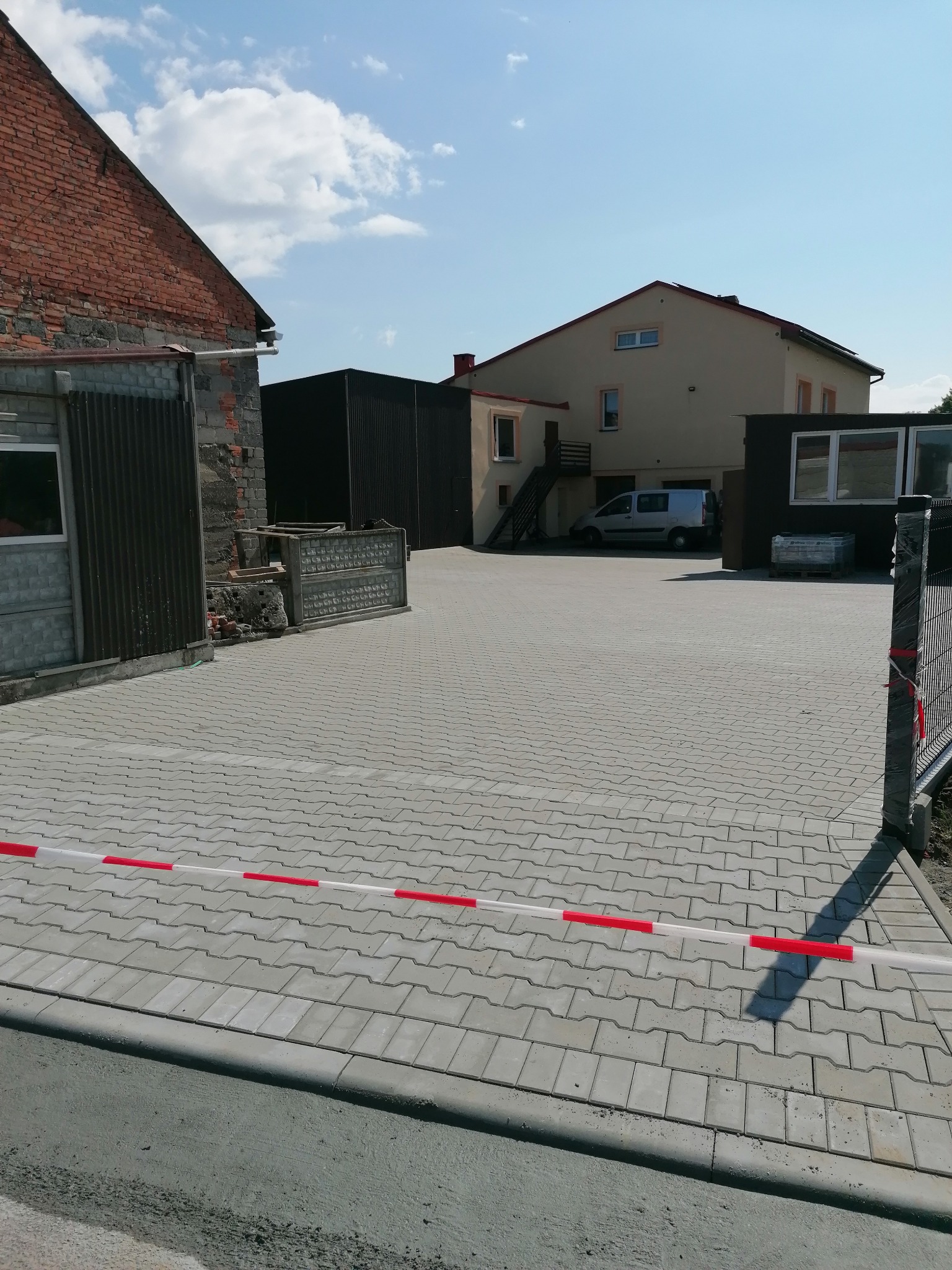 Szara kostka brukowa ułożona na posesji, częściowo odgrodzona taśmą ostrzegawczą. W tle budynki i zaparkowany samochód dostawczy.