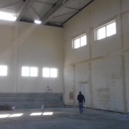 Firma Remontowo Budowlana Fachbud Piotr Grzegorzek - Wnętrze hali w stanie surowym, ściany częściowo przygotowane pod wykończenie, widoczne ślady po tynkowaniu, mężczyzna przechadza się po betonowej posadzce.