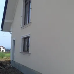 Elewacja domu jednorodzinnego po tynkowaniu, z widocznymi oknami z ozdobnymi ramami i wykończonym cokołem, na tle fragmentu ogrodu i sąsiednich budynk&oacute;w.