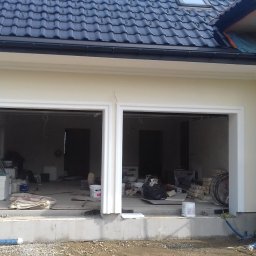 Firma Remontowo Budowlana Fachbud Piotr Grzegorzek - Elewacja budynku z dwoma otworami garażowymi w trakcie prac wykończeniowych, widoczne rusztowania i materiały budowlane wewnątrz.