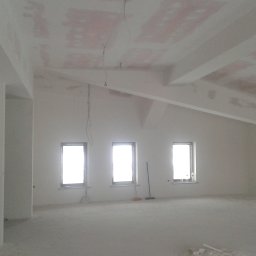 Firma Remontowo Budowlana Fachbud Piotr Grzegorzek - Wnętrze w trakcie remontu: surowe ściany po tynkowaniu, widoczne ślady po łatach na suficie, trzy okna z widokiem na zewnątrz, przewody elektryczne zwisające z sufitu, oraz szczotka oparta o ścianę.