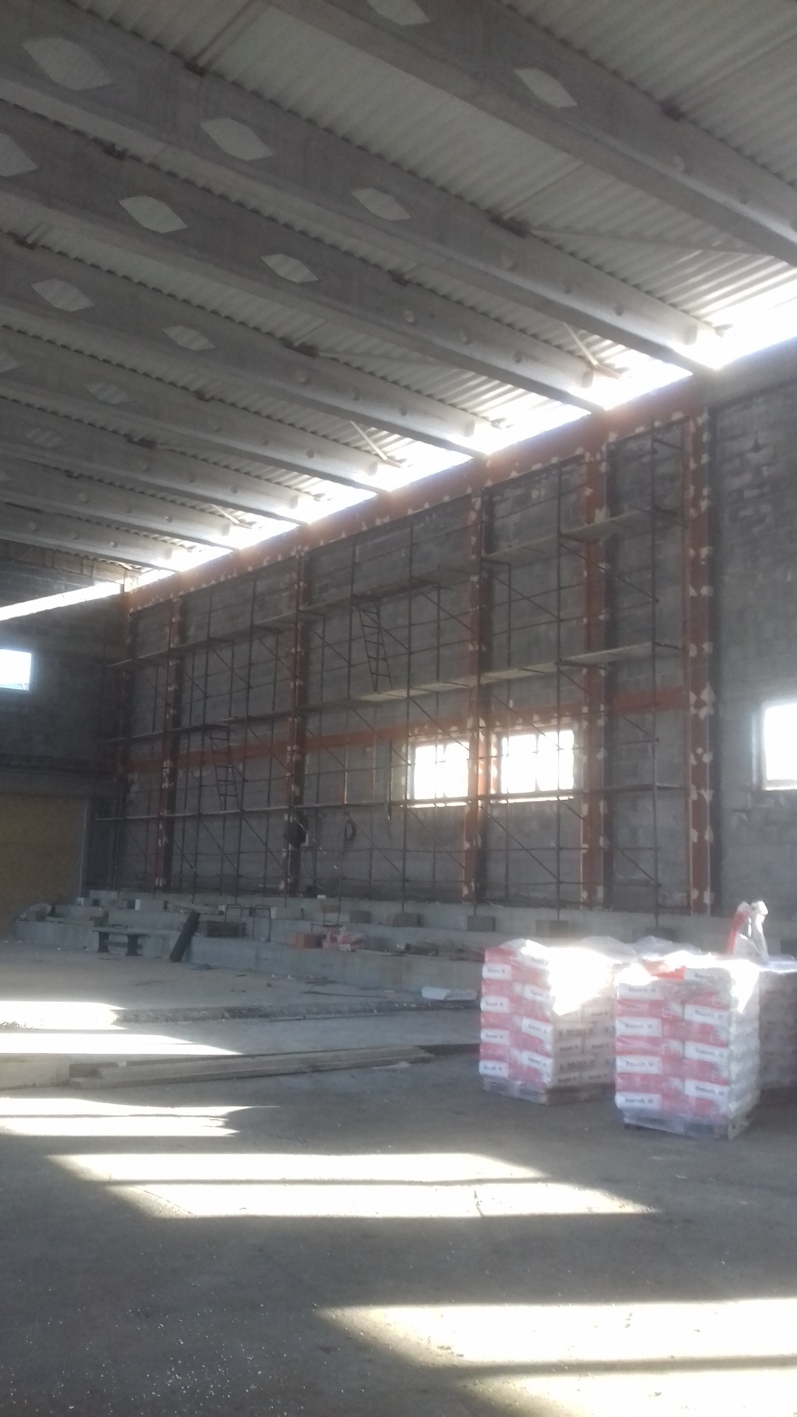 Wnętrze hali w trakcie prac budowlanych, widoczne rusztowania przy ścianie i palety z materiałami budowlanymi w tle, oświetlone wpadającym światłem słonecznym.