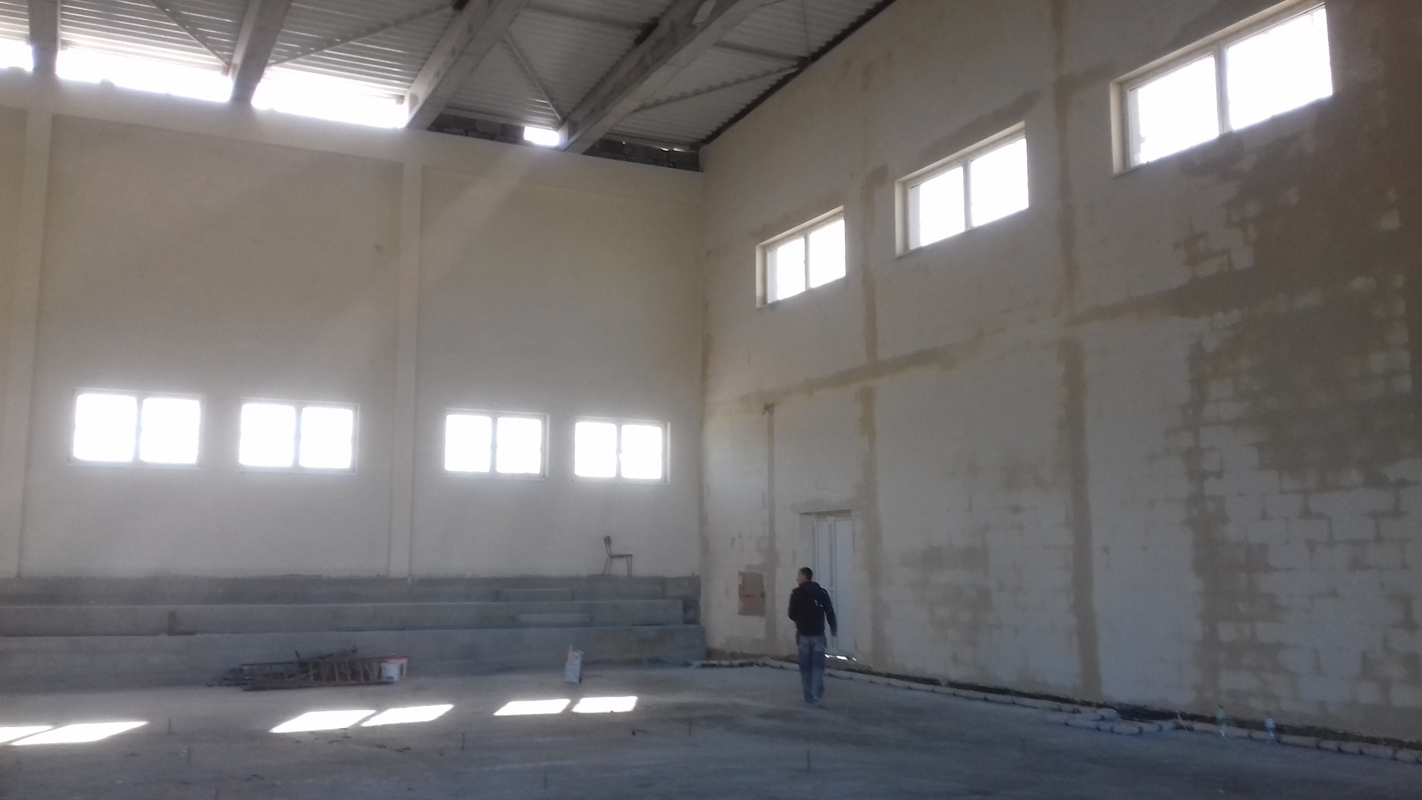 Wnętrze hali w stanie surowym, ściany częściowo przygotowane pod wykończenie, widoczne ślady po tynkowaniu, mężczyzna przechadza się po betonowej posadzce.