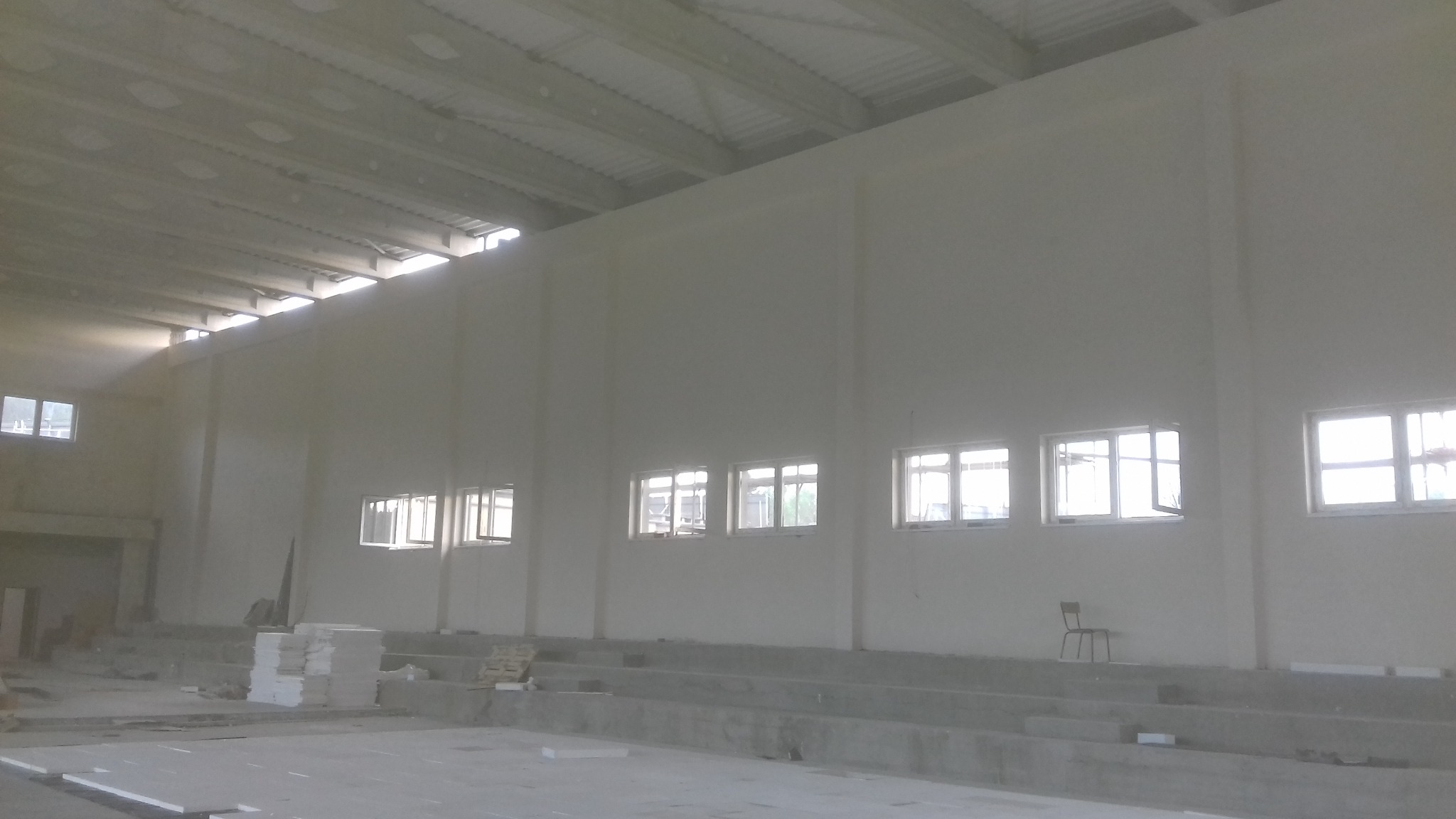 Wnętrze hali sportowej w trakcie prac wykończeniowych, widoczne gładkie, pomalowane na biało ściany, okna z widokiem na zewnątrz, surowy beton na podłodze i konstrukcja dachu, krzesło przy ścianie.