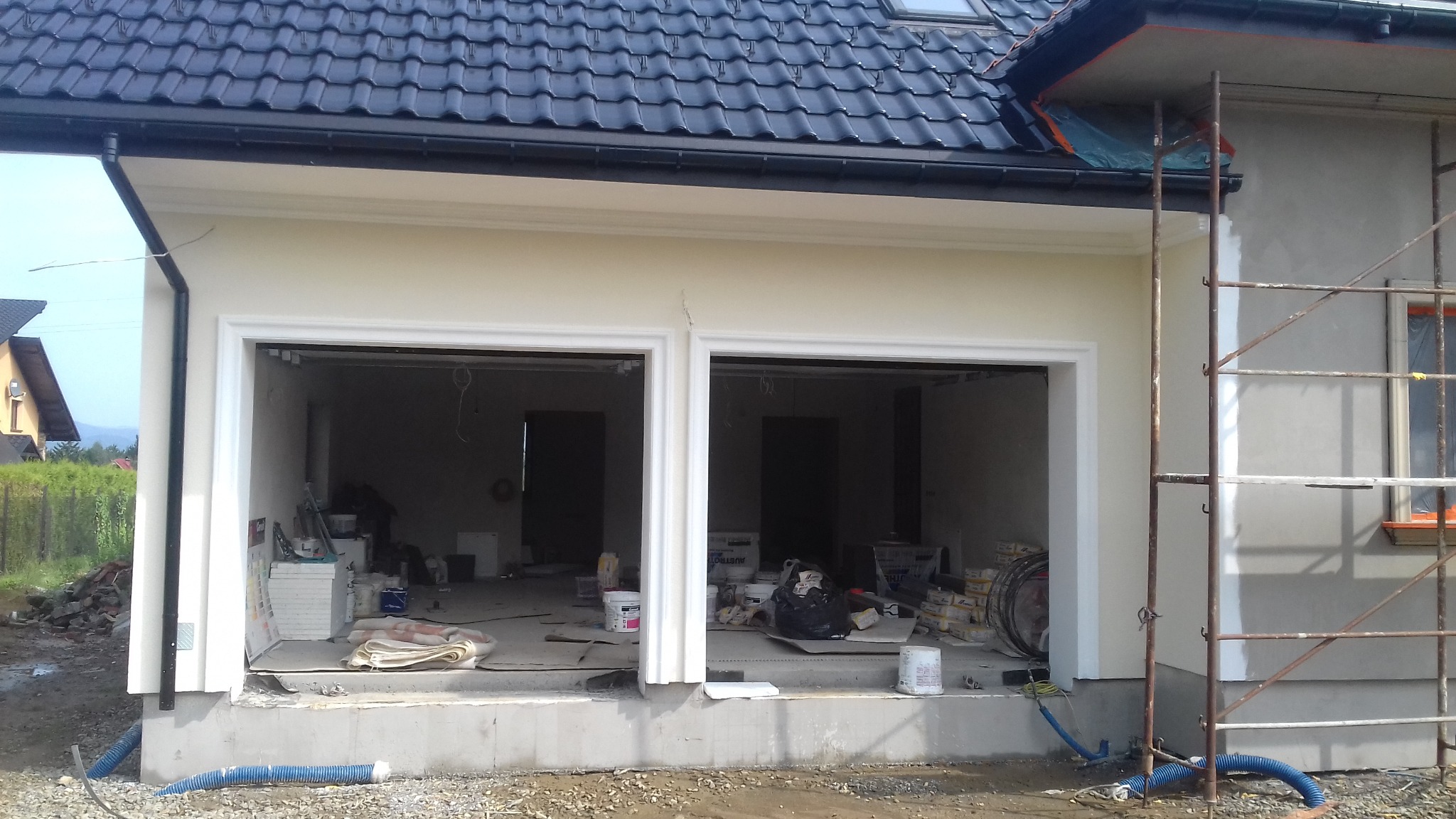 Elewacja budynku z dwoma otworami garażowymi w trakcie prac wykończeniowych, widoczne rusztowania i materiały budowlane wewnątrz.