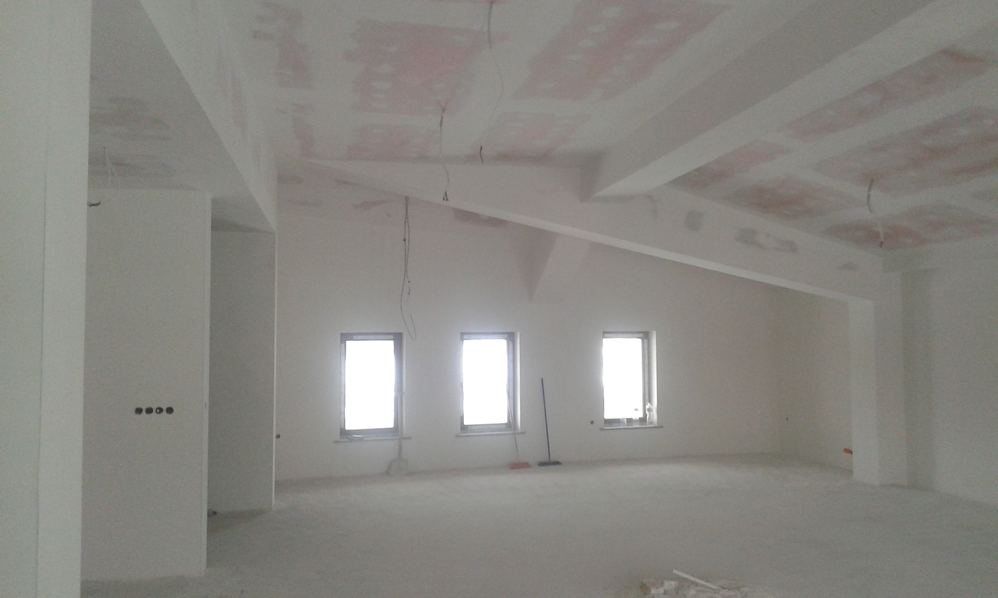 Wnętrze w trakcie remontu: surowe ściany po tynkowaniu, widoczne ślady po łatach na suficie, trzy okna z widokiem na zewnątrz, przewody elektryczne zwisające z sufitu, oraz szczotka oparta o ścianę.