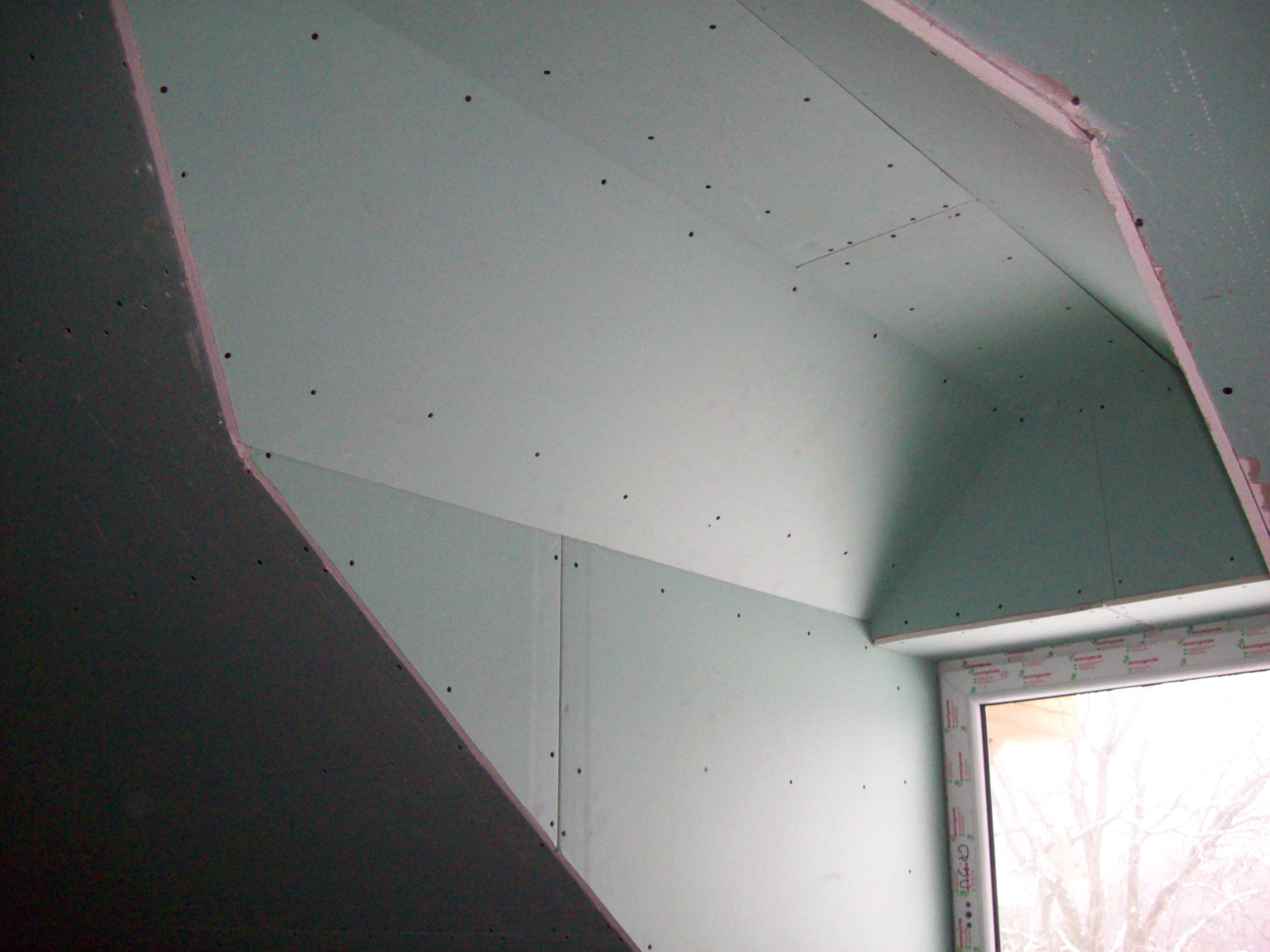 Montaż płyt gipsowo-kartonowych na poddaszu, widoczne wkręty mocujące i okno z folią ochronną.