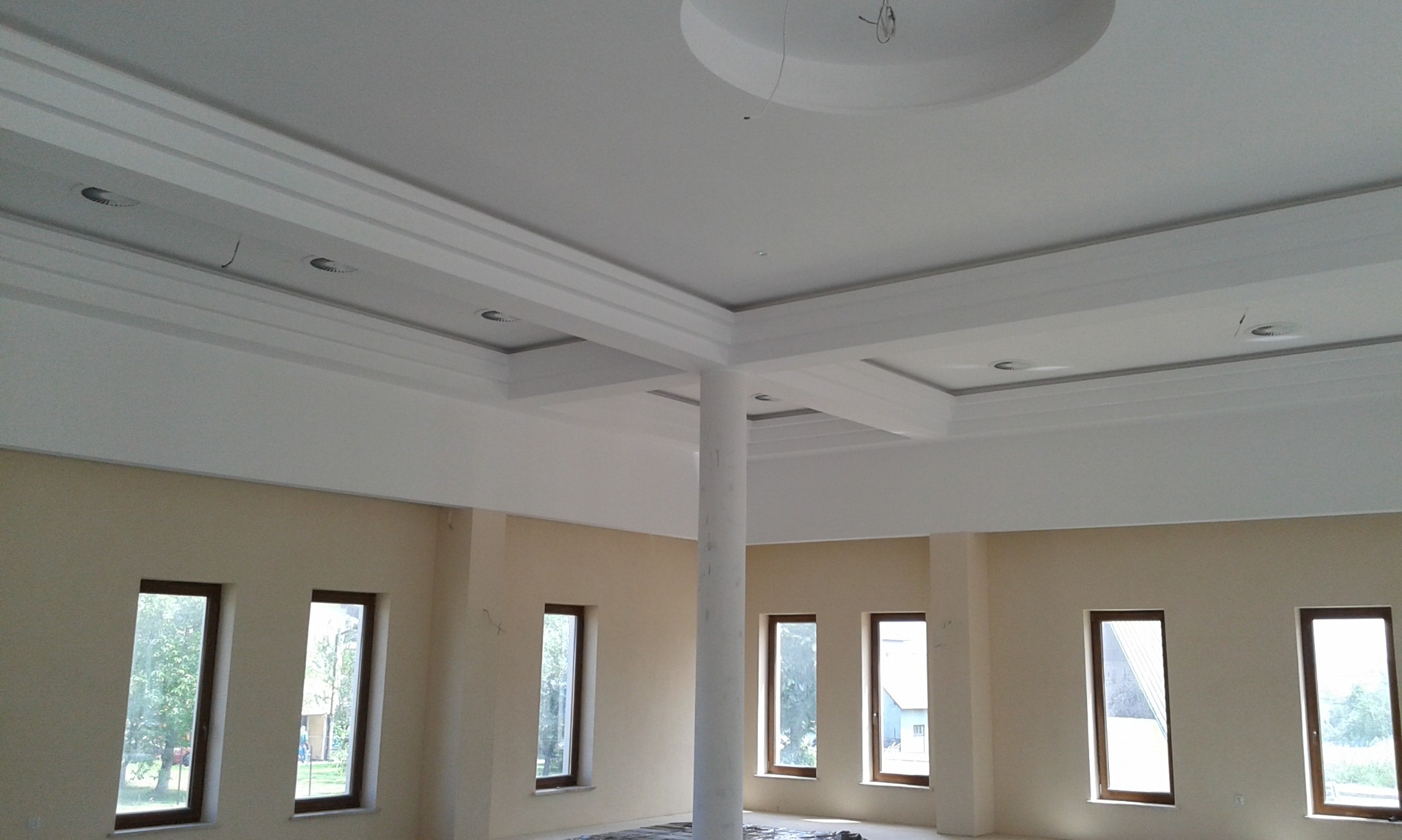 Wnętrze dużego pokoju po remoncie z kolumną nośną na środku, widoczne okna z widokiem na zieleń, gładkie ściany w odcieniach beżu oraz biały sufit z dekoracyjnymi elementami i oświetleniem punktowym.