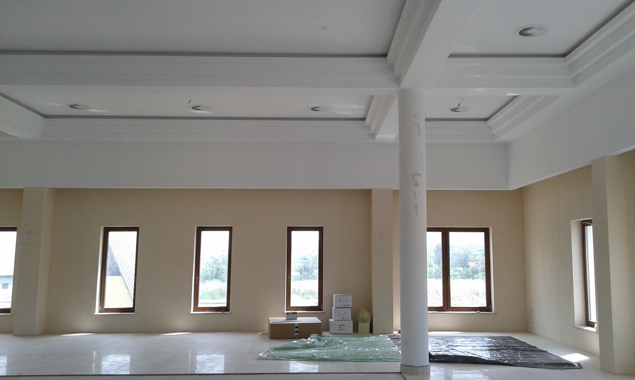 Wnętrze w stanie deweloperskim z wykończonymi ścianami w odcieniach beżu, białym sufitem z ozdobnymi elementami i kolumną, zabezpieczone podłogi folią malarską, widoczne kartony i puszka farby.