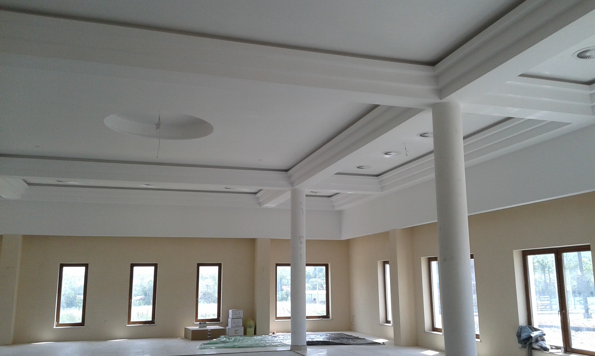 Wnętrze przestronnego pomieszczenia z kolumnami i dekoracyjnym sufitem, ściany częściowo pomalowane na beżowo, widoczne okna i zabezpieczona folią podłoga.