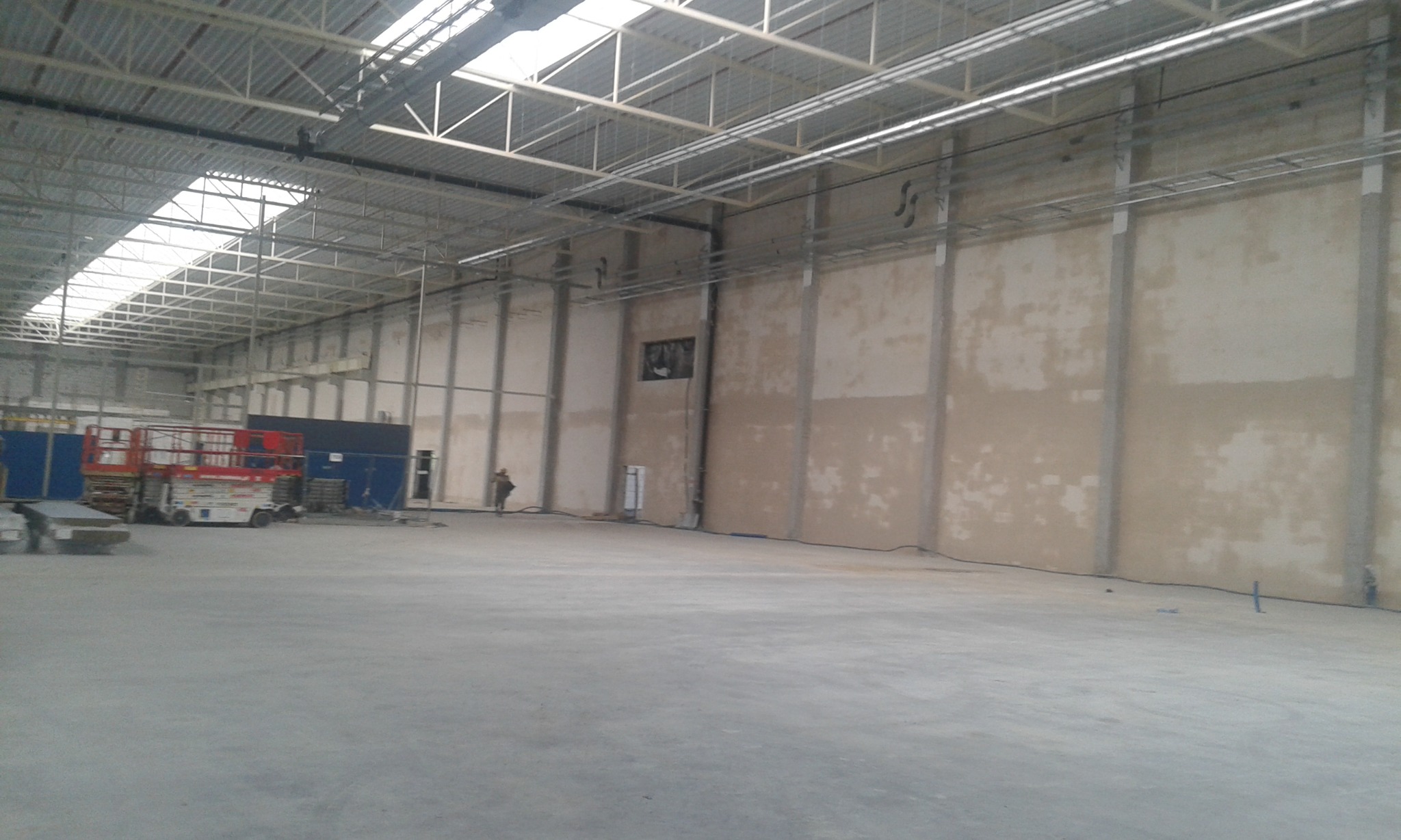 Wnętrze hali przemysłowej w trakcie prac wykończeniowych, widoczne surowe ściany z fragmentami nałożonego tynku, metalowa konstrukcja dachu i podnośnik koszowy w tle.