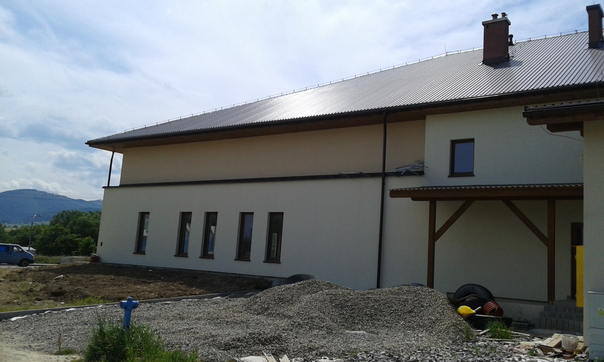 Jasna elewacja budynku z ciemną dachówką w Cisiecu, widoczny stos kruszywa na pierwszym planie, częściowo zagospodarowany teren.