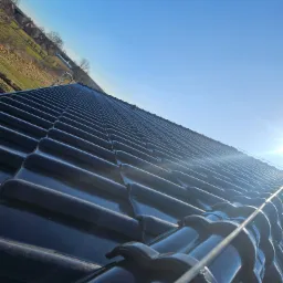 Błyszczący, nowy dach z ciemnej dachówki ceramicznej pod błękitnym niebem, widok z dołu pod kątem. W tle zieleń i budynki.