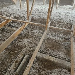 IzoProDach Kamil Nowak - Poddasze z widoczną konstrukcją drewnianą i warstwą izolacji z wełny mineralnej, przewody elektryczne, rury wentylacyjne. Ocieplenie wykonane w szarym odcieniu.