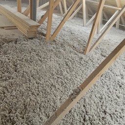 IzoProDach Kamil Nowak - Poddasze z drewnianą konstrukcją wypełnione szarą wełną izolacyjną. Widoczny fragment komina i deski. Nierównomierne oświetlenie podkreśla strukturę materiału.