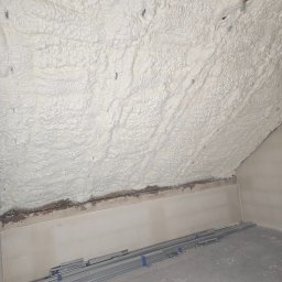 IzoProDach Kamil Nowak - Poddasze po natrysku pianki PUR, widoczne metalowe profile na podłodze. Biała, nierówna struktura pianki szczelnie wypełnia przestrzeń.