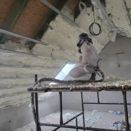 IzoProDach Kamil Nowak - Pracownik w kombinezonie aplikuje pianę PUR na poddaszu, stojąc na rusztowaniu. Widoczne ocieplone elementy konstrukcji dachu i okno dachowe. Detal: przewody i pianka na rusztowaniu.