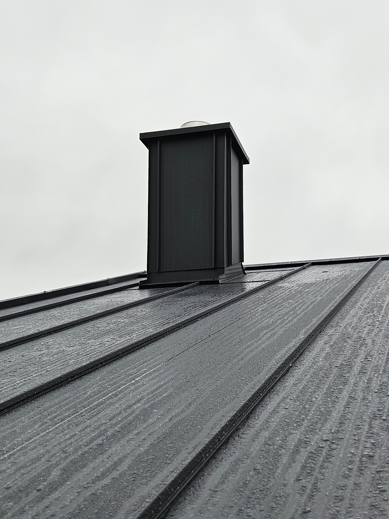 Szary, metalowy dach z kominem w kształcie prostopadłościanu, widziany z dołu pod kątem, w pochmurny dzień. Widoczne detale łączeń blachy dachowej.