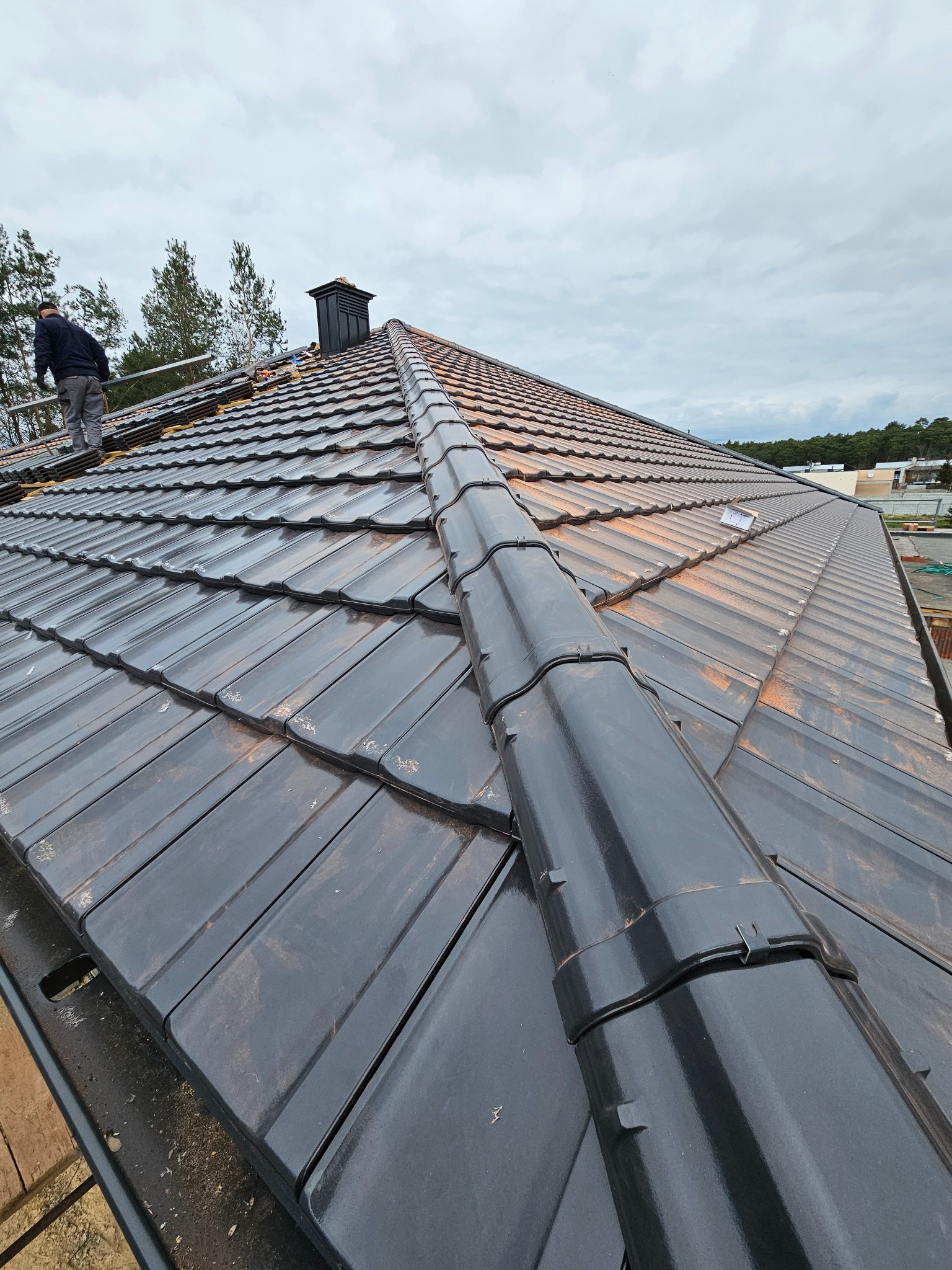 Układanie ciemnej dachówki na skośnym dachu domu jednorodzinnego, widok z dołu z pracownikiem na drabinie, komin w tle, pochmurne niebo.