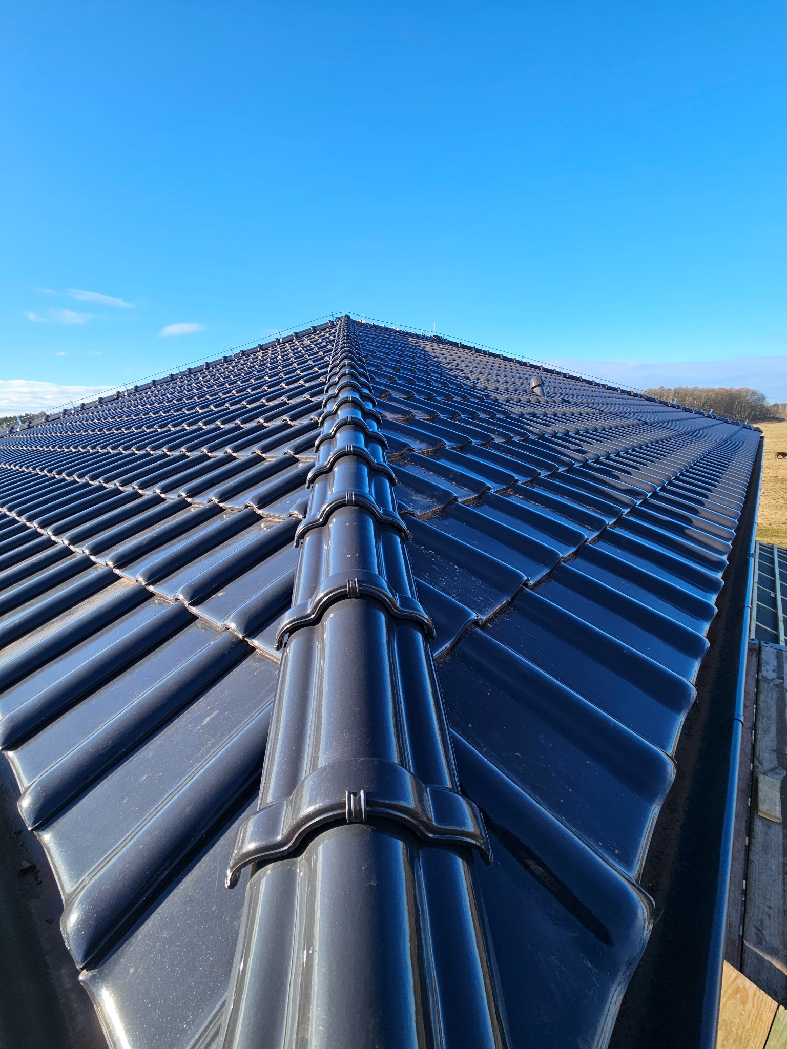 Błyszczący, ciemny dach z dachówki ceramicznej w pełnym słońcu, widziany z góry. Na tle błękitne niebo i fragment krajobrazu.