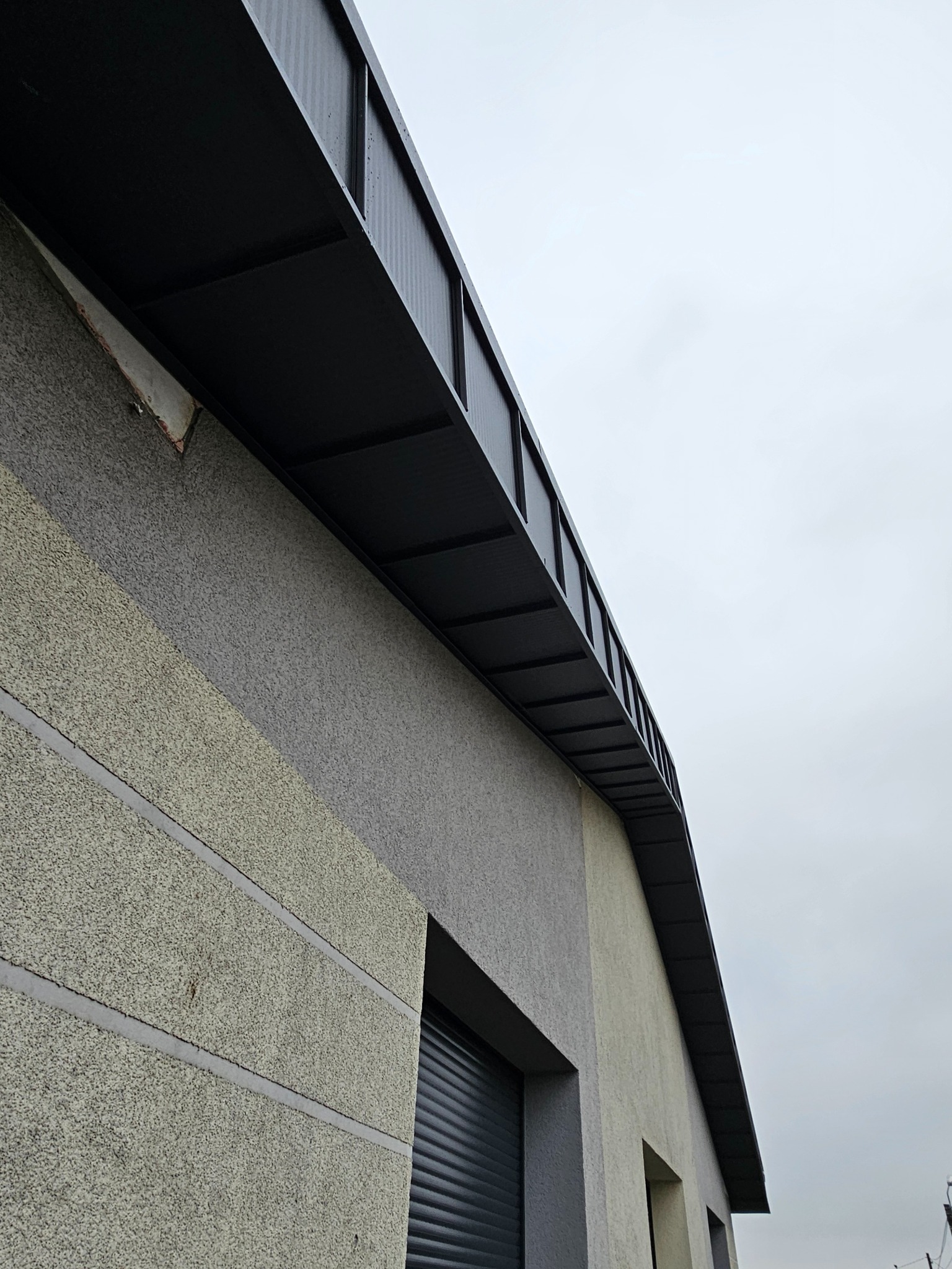 Szary budynek z oknami i czarnym, metalowym okapem w pochmurny dzień. Widok z dołu, ukazujący strukturę elewacji i detale wykończenia okapu.