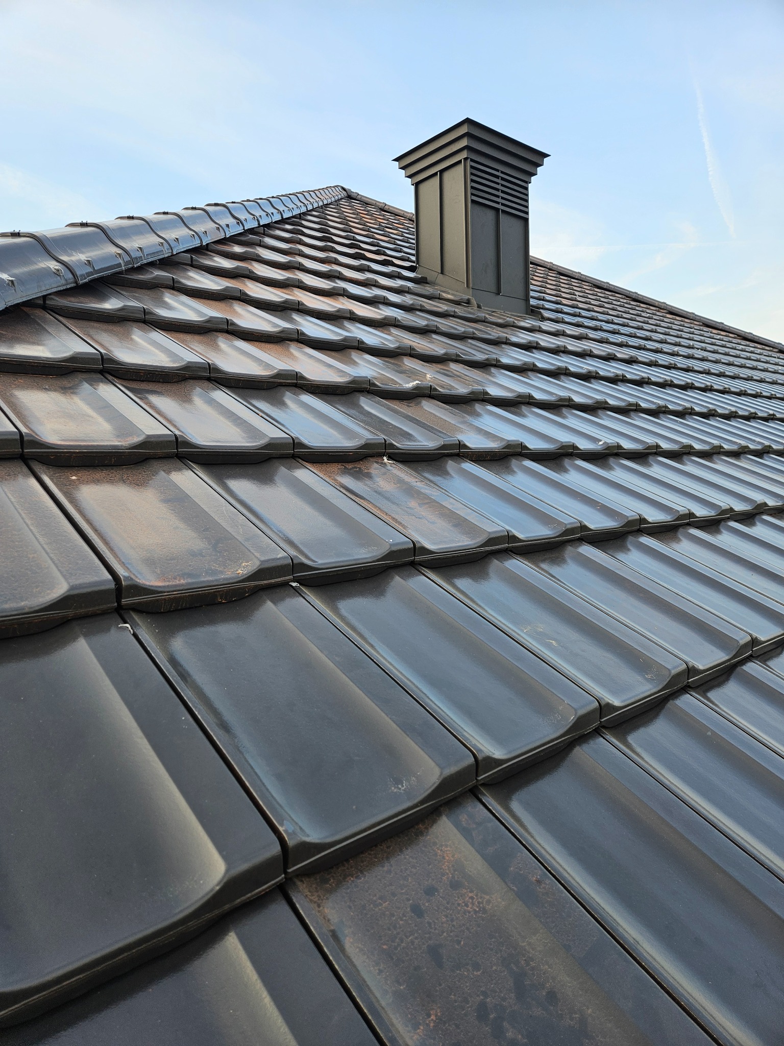 Błyszczący, grafitowy dach z ceramicznej dachówki ułożonej w rzędach, z kominem w kolorze dachu, na tle błękitnego nieba z delikatnymi smugami.