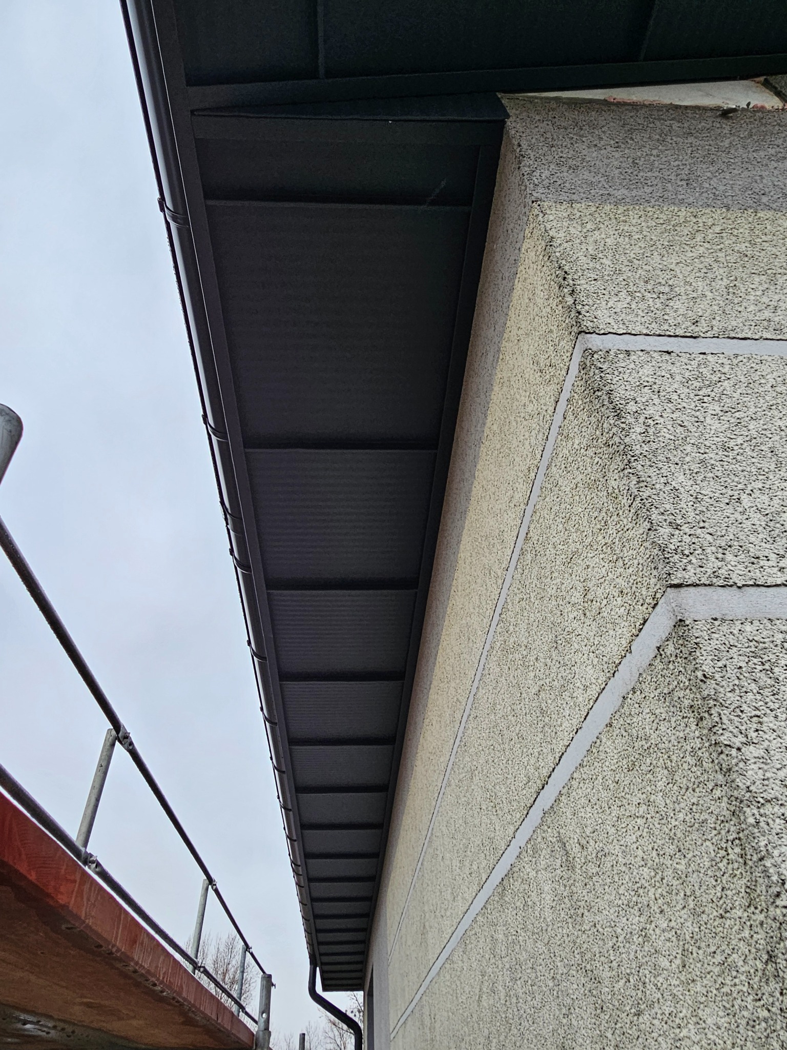 Poddasze po ociepleniu, widoczne czarne panele izolacyjne pod okapem. Elewacja budynku z fakturą 'baranka' i szare listwy dekoracyjne. Fragment rusztowania.