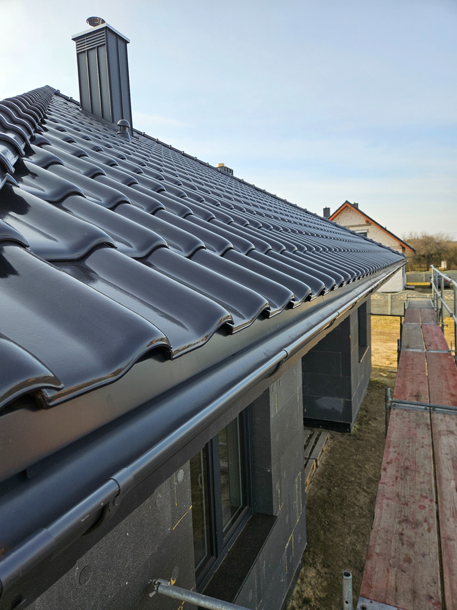 Nowoczesny, czarny dach z połyskującą dachówką ceramiczną i systemem rynnowym. Budynek w trakcie budowy, ocieplony styropianem grafitowym, widoczne rusztowania.