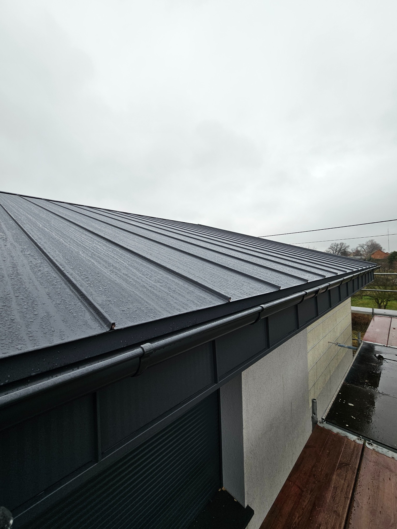 Metalowy dach z orynnowaniem w trakcie montażu, widok z góry, w tle pochmurne niebo. Elementy rusztowania.