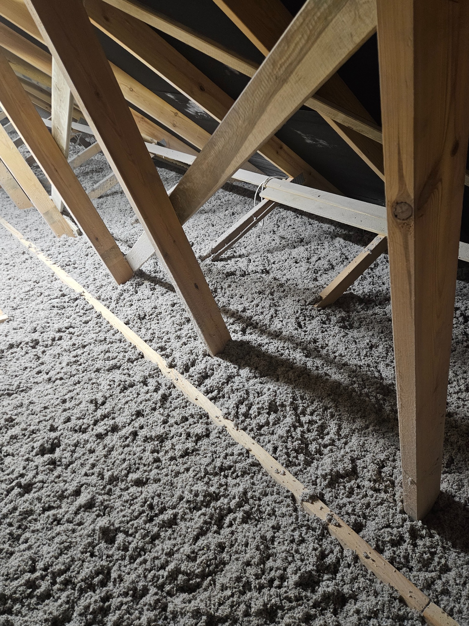 Szara izolacja z celulozy wypełnia przestrzeń między drewnianymi krokwiami poddasza, tworząc gęstą warstwę izolacyjną.