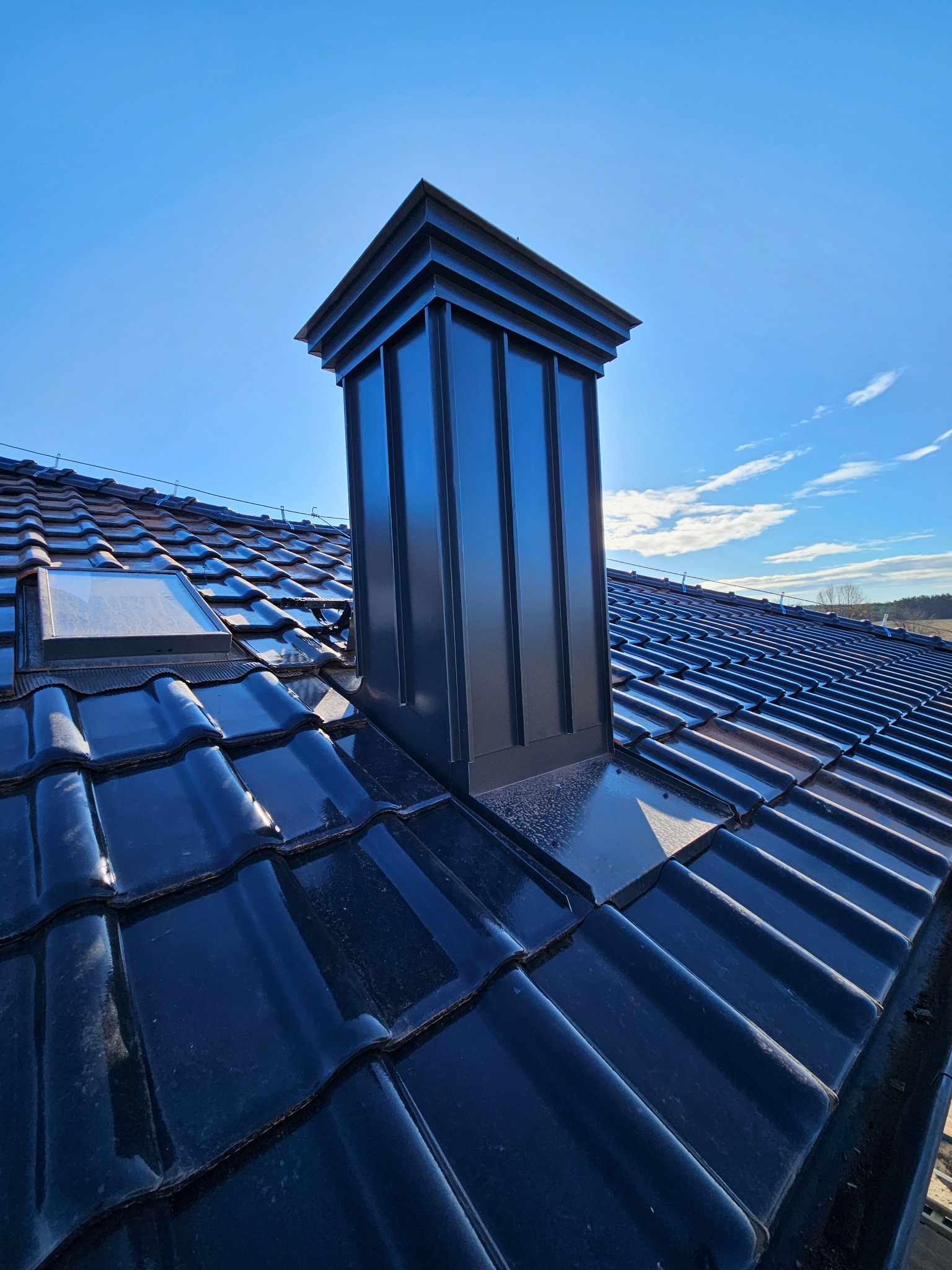 Błyszcząca, czarna dachówka ceramiczna z oknem dachowym i nowoczesnym, metalowym kominem na tle błękitnego nieba z delikatnymi obłokami. Widok z dołu.