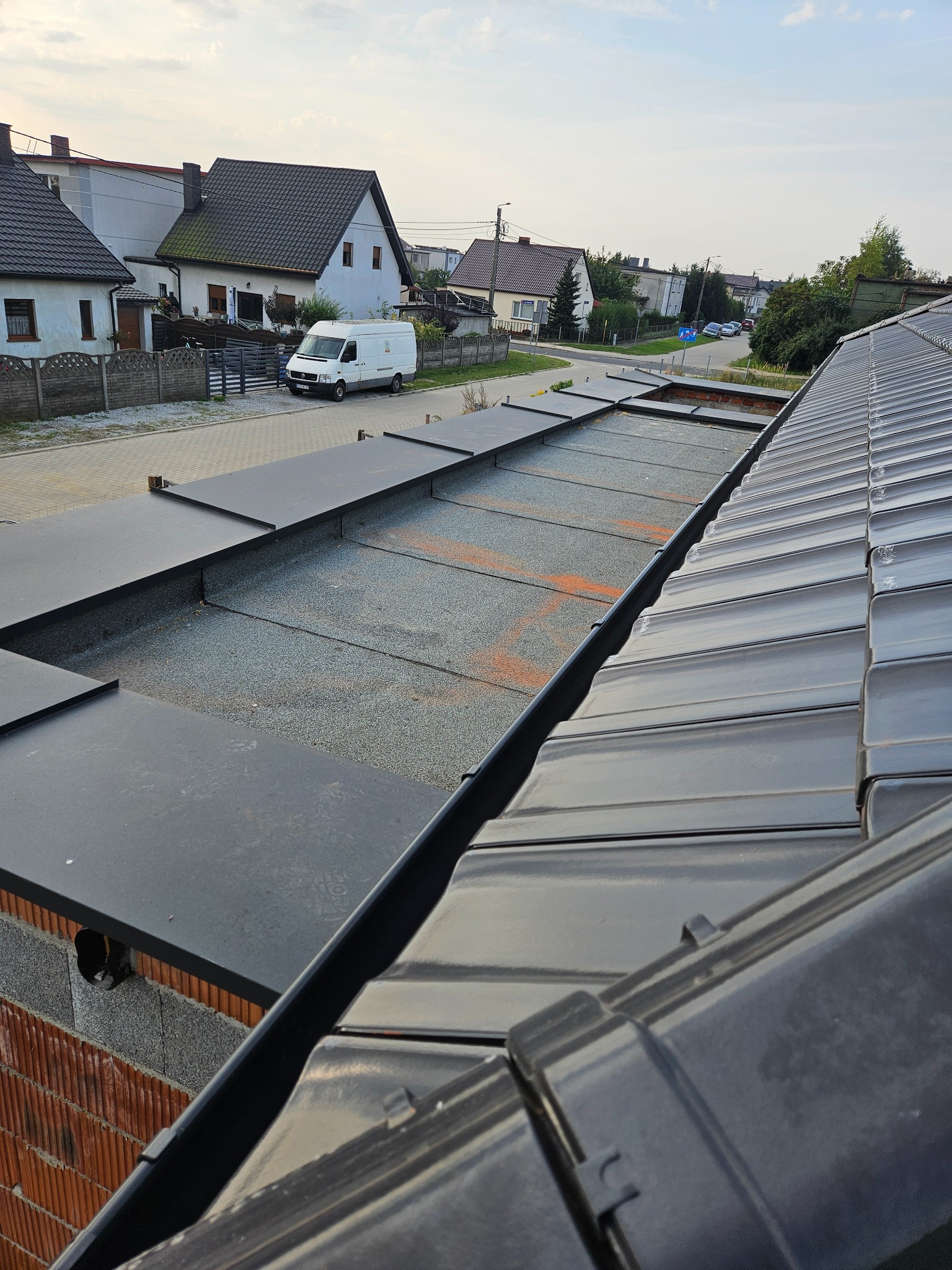 Widok z góry na fragment dachu z czarną blachodachówką i płaską powierzchnią pokrytą szarym granulatem. W tle zabudowa jednorodzinna i błękitne niebo.