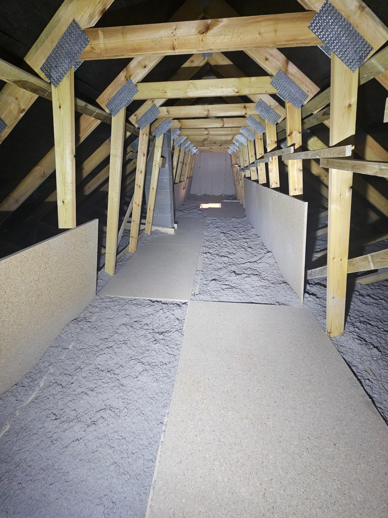 Ocieplone poddasze wełną mineralną, widok z perspektywy. Elementy drewnianej konstrukcji dachu z metalowymi wzmocnieniami. Fragment podłogi wyłożony płytami OSB.