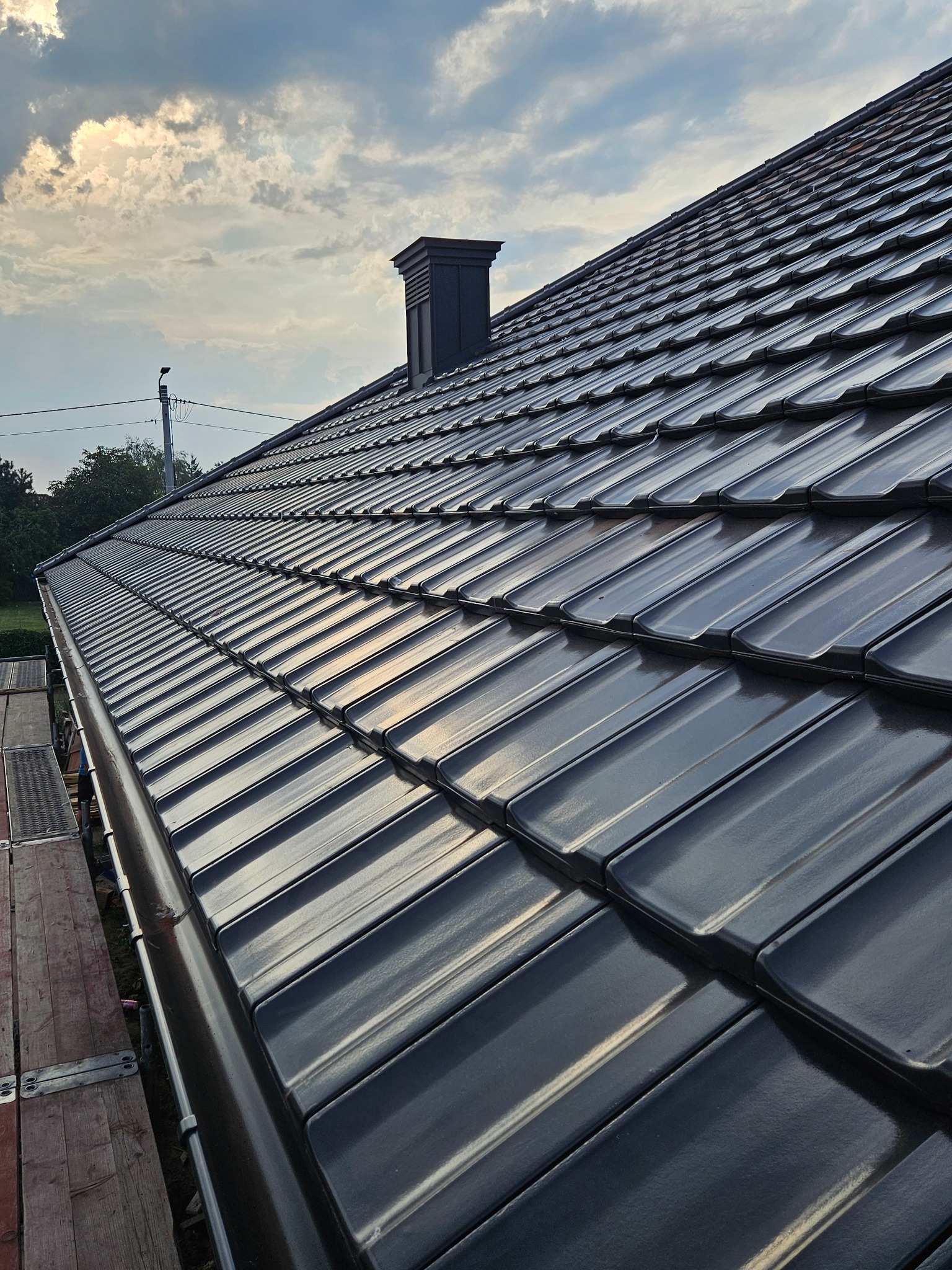 Błyszczący, grafitowy dach z kominem w perspektywie, widok z rusztowania. Tło: zachmurzone niebo. Układanie dachówki etap końcowy.