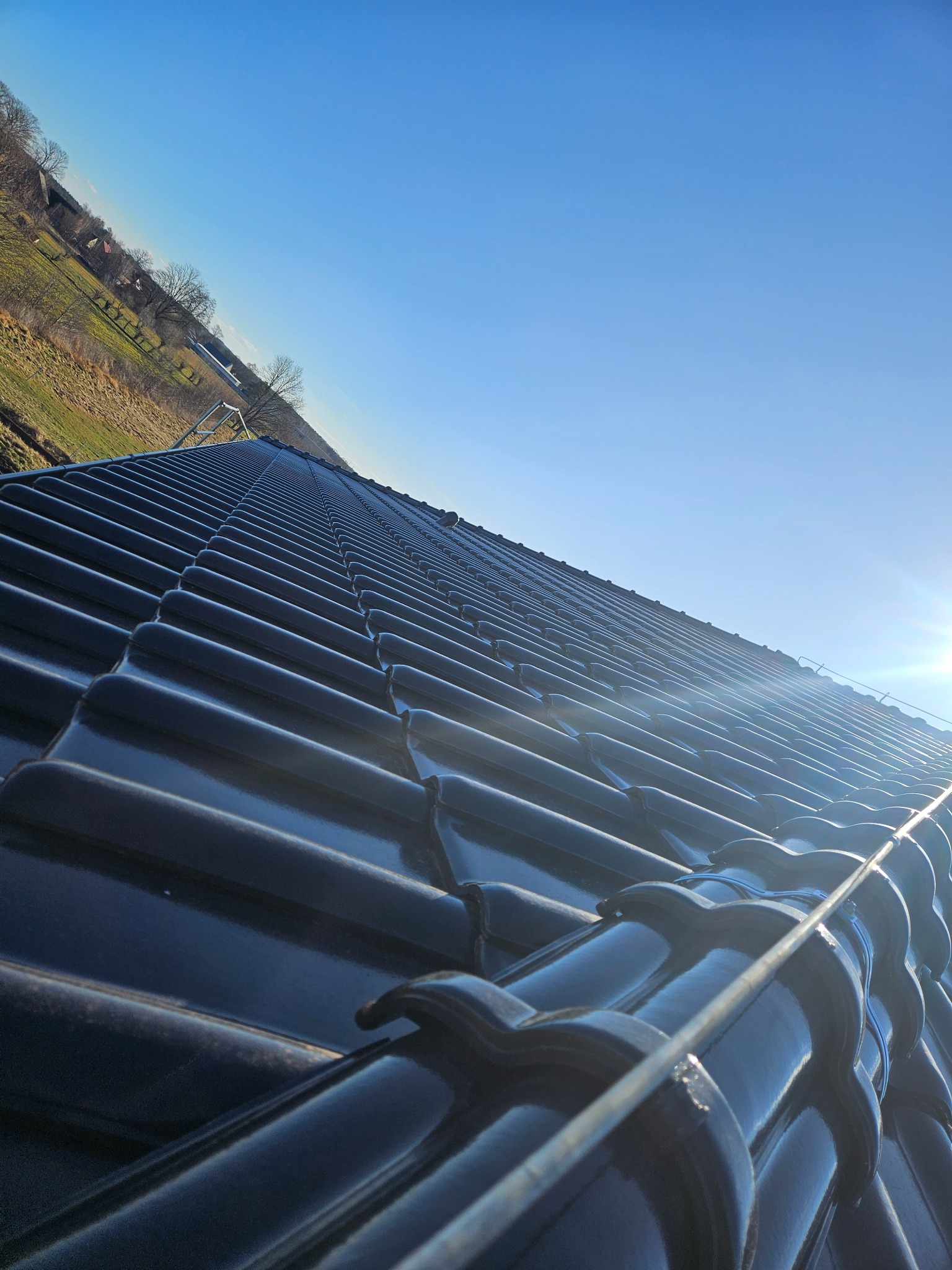 Błyszczący, nowy dach z ciemnej dachówki ceramicznej pod błękitnym niebem, widok z dołu pod kątem. W tle zieleń i budynki.