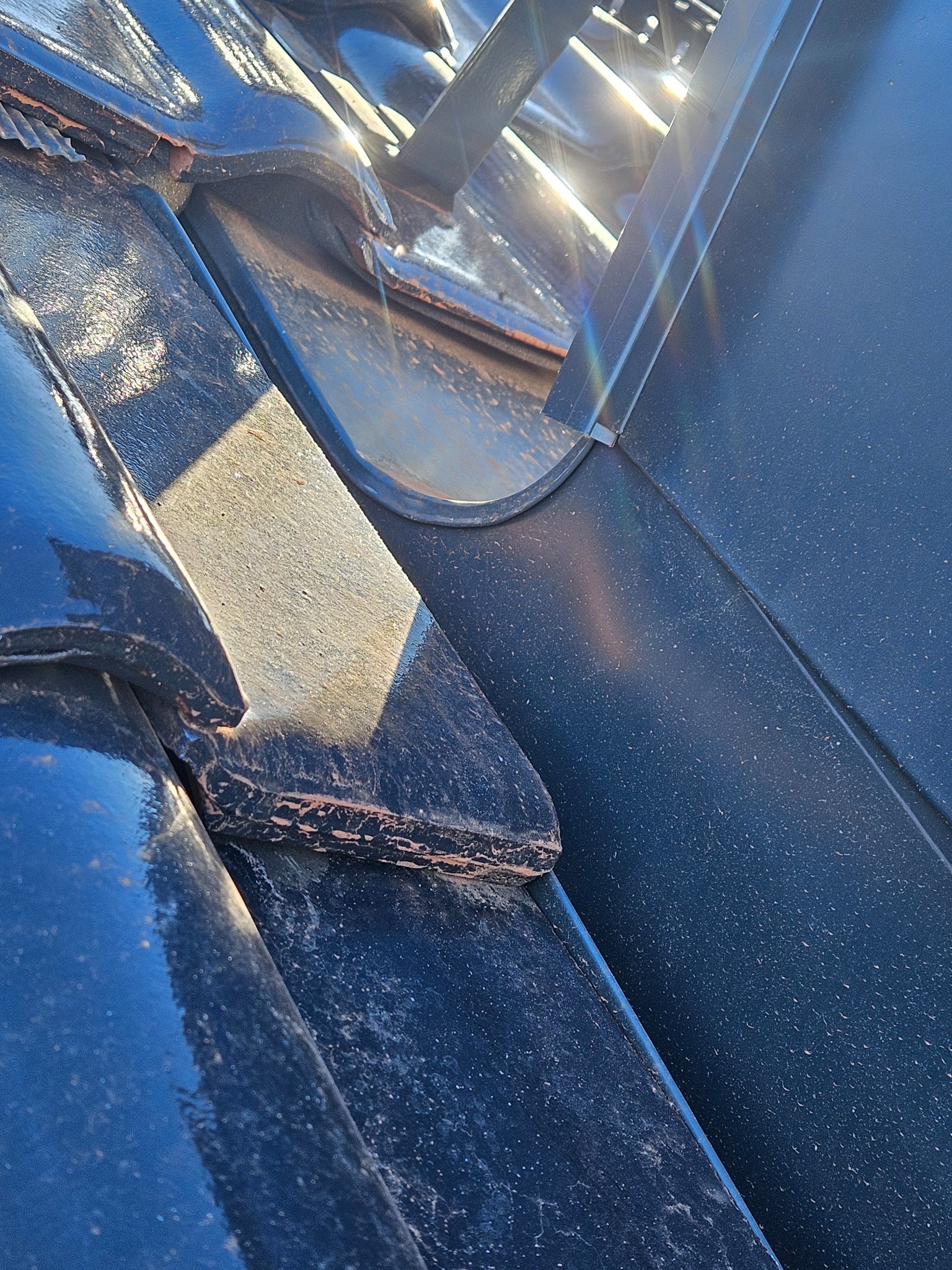 Szczegółowe ujęcie ciemnych dachówek z metalowym elementem w słoneczny dzień. Widoczne detale faktury i połączenia materiałów.