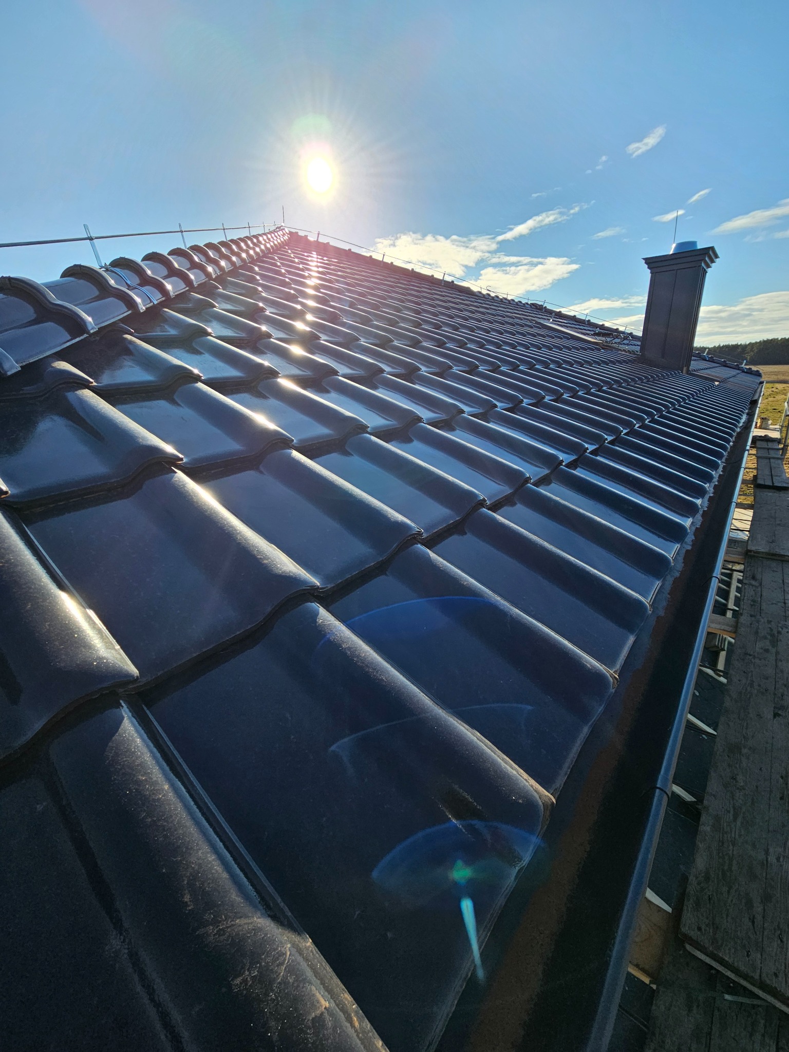 Połyskująca, ciemna dachówka ceramiczna na ukończonym dachu domu w słoneczny dzień. Widoczny komin i fragment rusztowania.