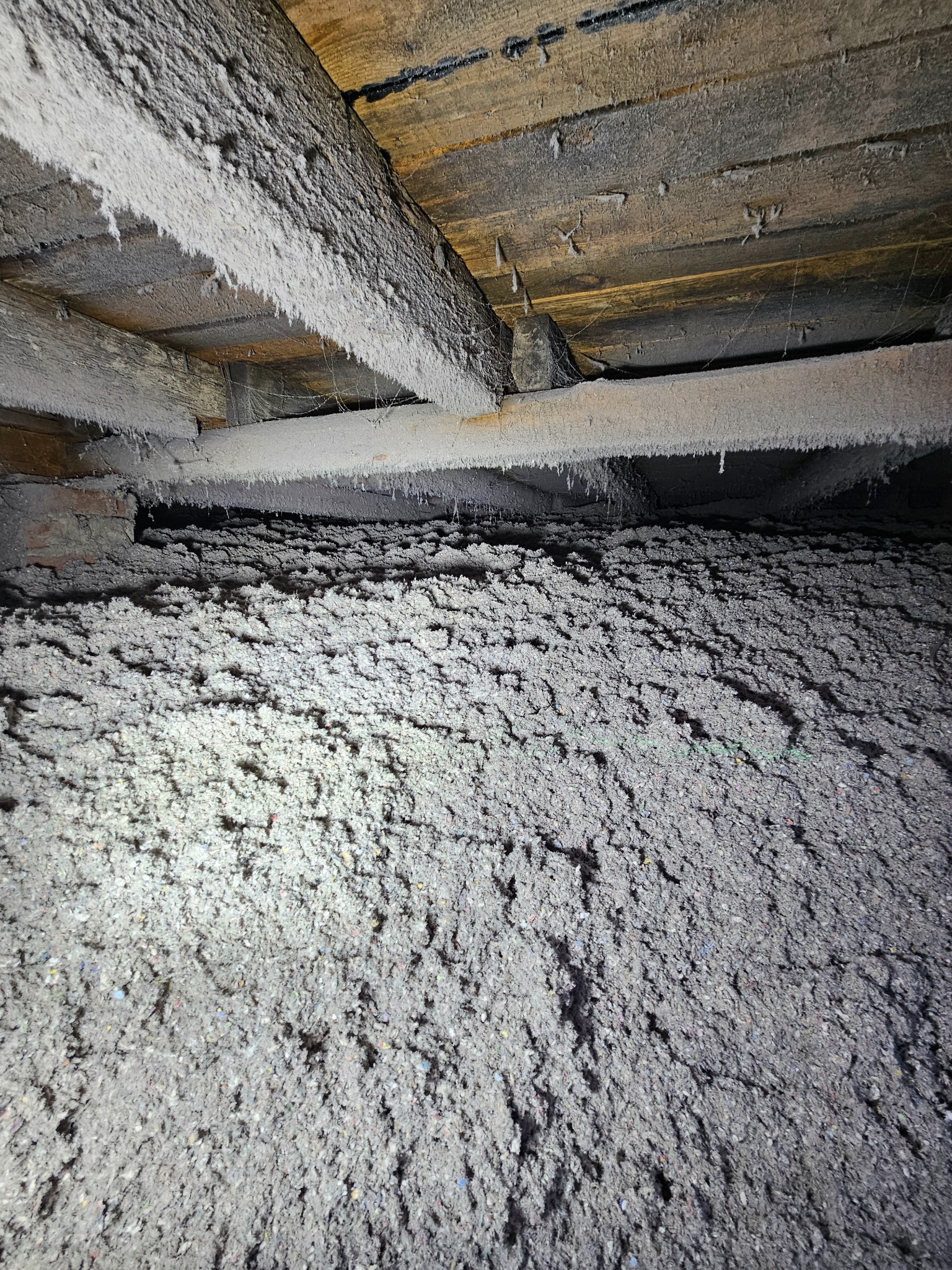 Szara warstwa izolacyjna z wełny mineralnej wypełnia przestrzeń pod drewnianą konstrukcją stropu poddasza, oświetlona punktowo. Widoczne drewniane belki.