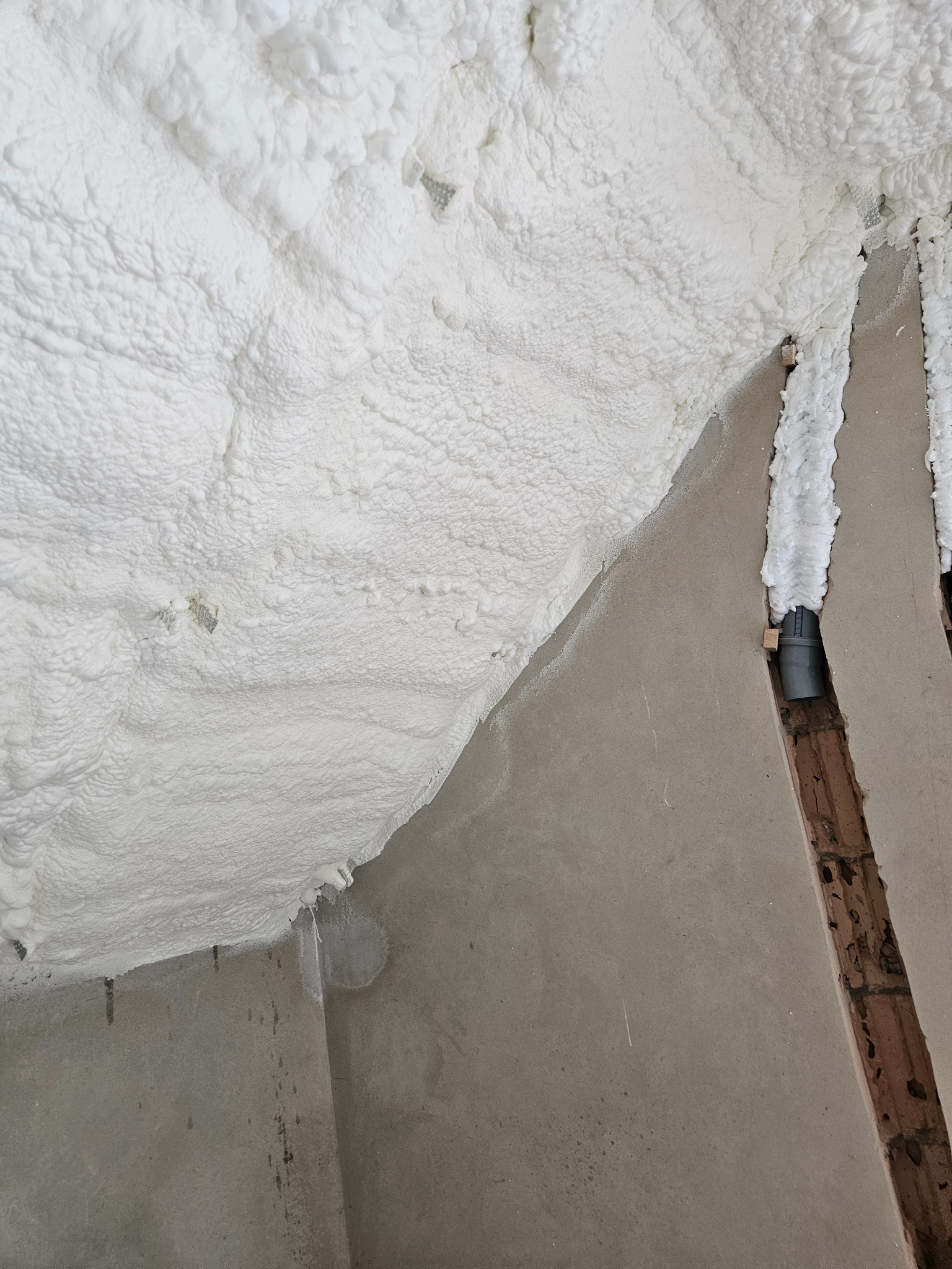 Poddasze ocieplone pianką PUR, widoczne drewniane elementy konstrukcyjne i szara ściana. Pianka pokrywa nierównomiernie powierzchnię, tworząc charakterystyczną strukturę.