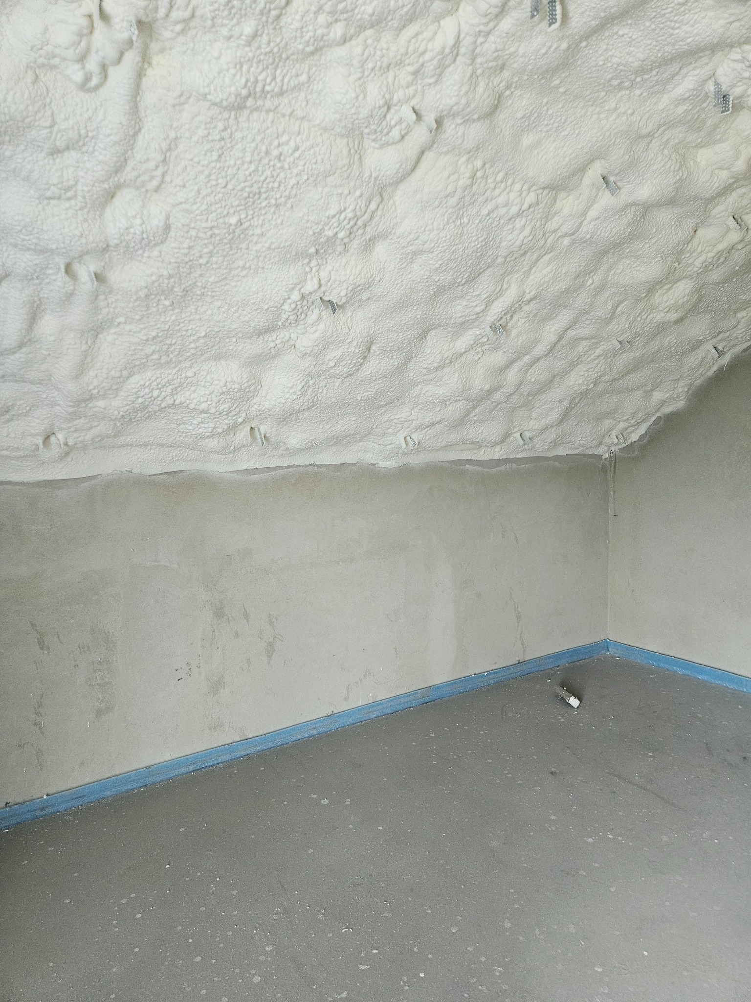 Poddasze po ociepleniu pianką PUR, widoczne metalowe uchwyty mocujące. Szara ściana i podłoga, zabezpieczone niebieską taśmą. Wykończony etap prac.