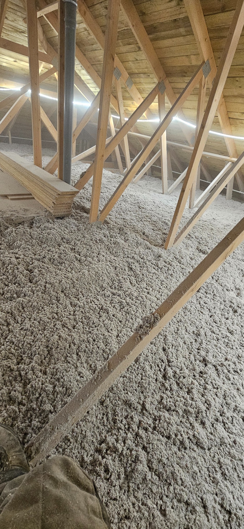 Poddasze z drewnianą konstrukcją wypełnione szarą wełną izolacyjną. Widoczny fragment komina i deski. Nierównomierne oświetlenie podkreśla strukturę materiału.