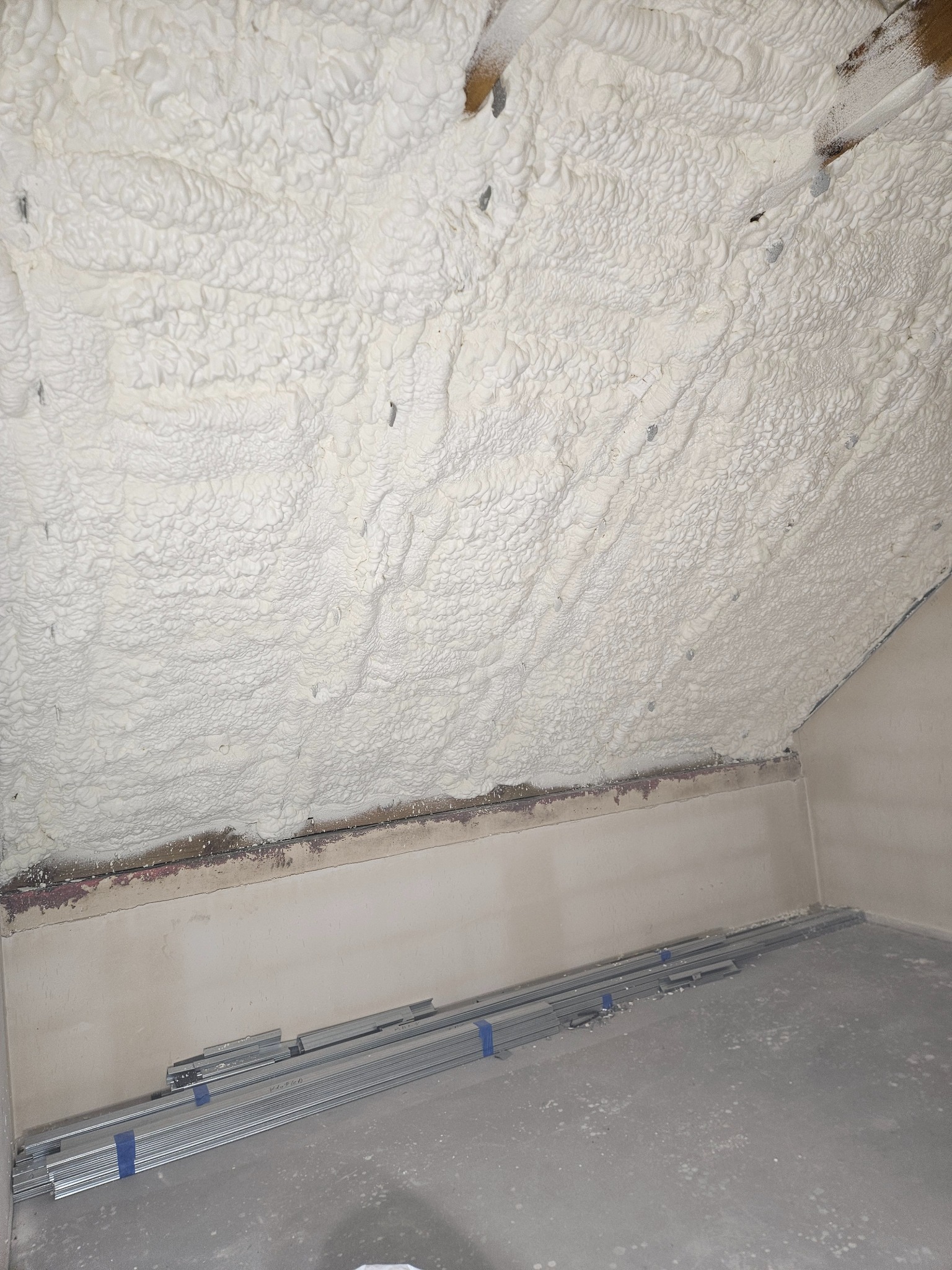 Poddasze po natrysku pianki PUR, widoczne metalowe profile na podłodze. Biała, nierówna struktura pianki szczelnie wypełnia przestrzeń.