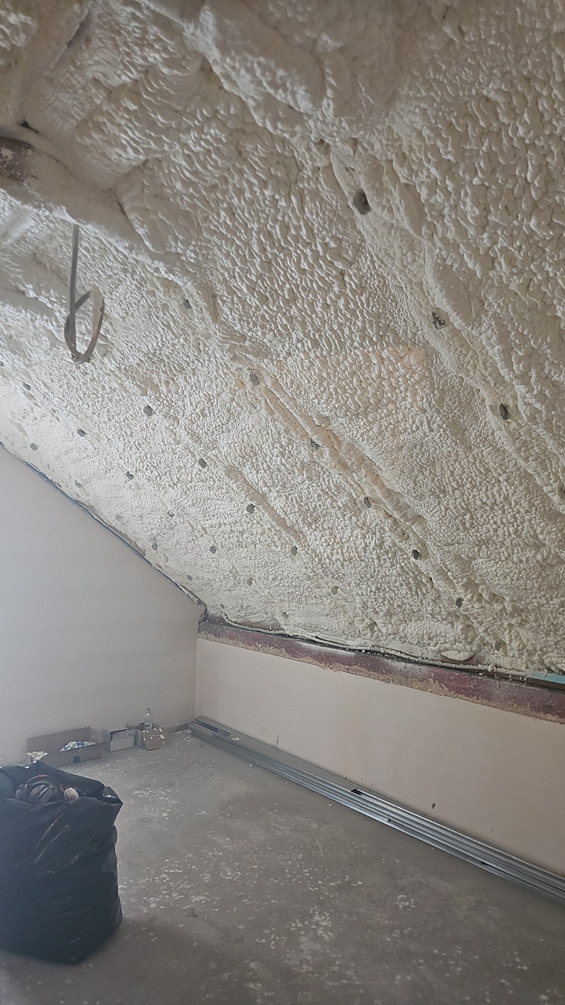 Poddasze po ociepleniu pianką PUR, widoczne równomierne pokrycie izolacją, elementy konstrukcyjne i ślady pianki na podłodze. Czarny worek na śmieci w rogu.